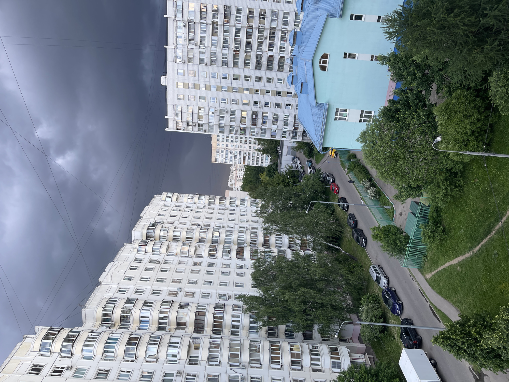 The image depicts an urban scene, likely in a residential area of Moscow, Russia, given the architectural style of the buildings. The view is from an elevated position, possibly an upper floor of a building.
The scene is dominated by two large, white apartment buildings with numerous windows. One building has a unique, stepped architectural design. A light blue building with a dark blue roof sits adjacent to one of the apartment buildings.
A street runs between the buildings, lined with trees and parked cars. A sidewalk is visible alongside the street. The sky is overcast and dark, suggesting an approaching storm. Power lines are visible crossing the sky. There are no people visible in the image.