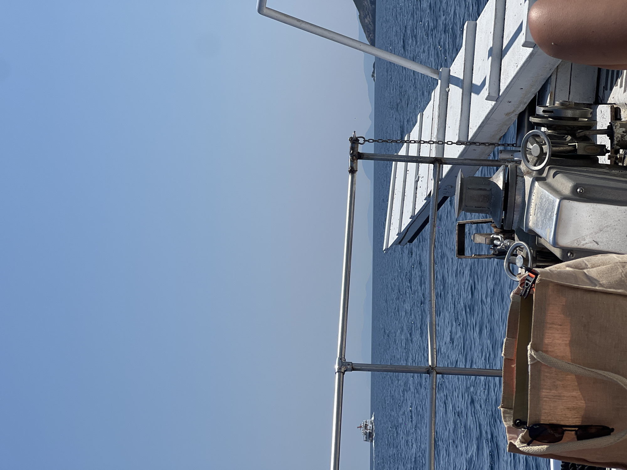 The image depicts a view from the deck of a boat sailing on the Aegean Sea under a clear blue sky. In the distance, a ferry boat is visible on the horizon, and coastal hills or islands can be seen. The foreground shows part of the boat's deck, including metal railings, a white ramp-like structure, a winch mechanism, and a canvas tote bag with sunglasses. A person's leg is also visible in the lower right corner. The scene suggests a bright, sunny day at sea in Greece.