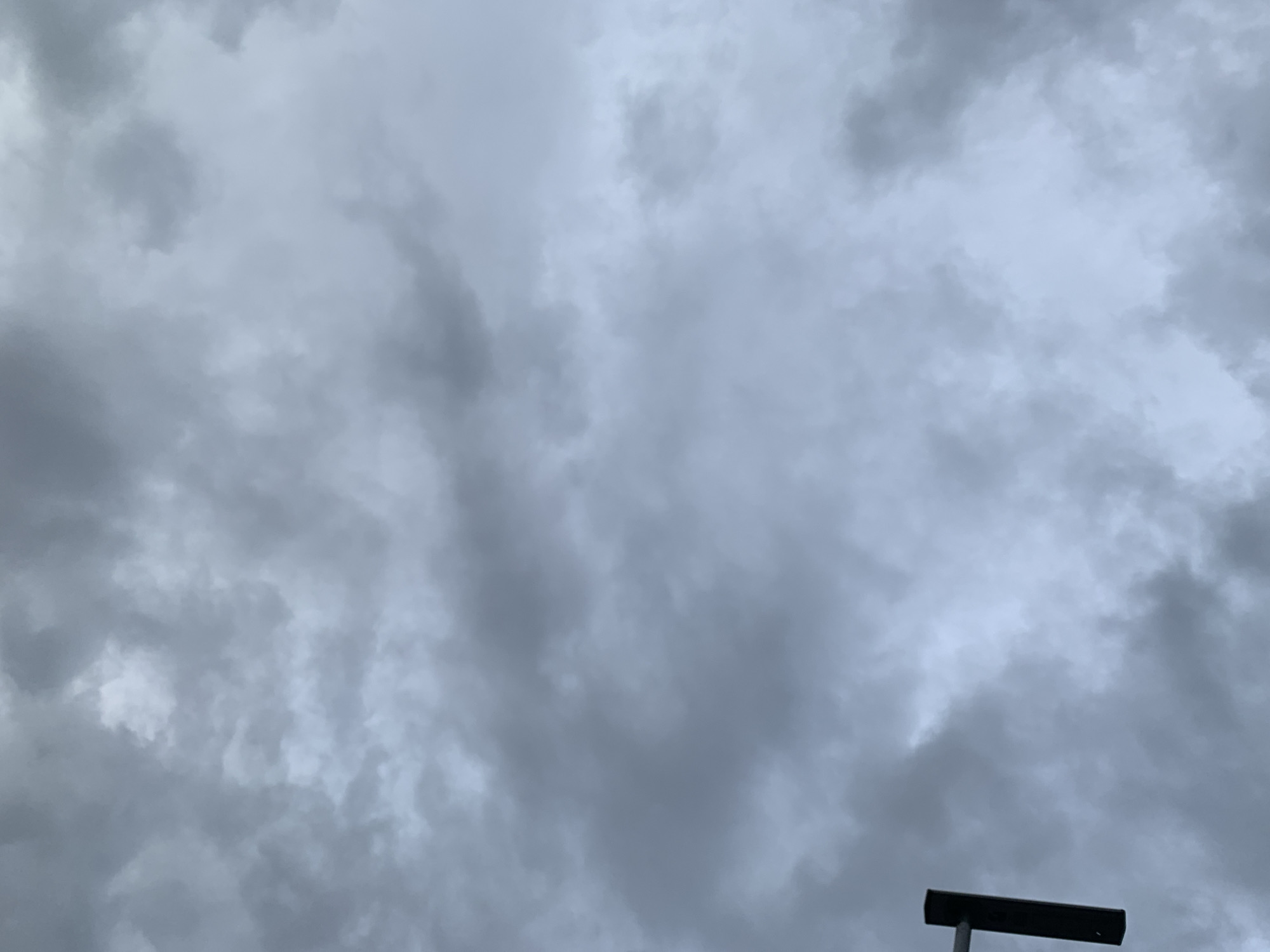 The image shows an overcast sky with dark, heavy clouds. The overall tone is dark and gloomy, suggesting a potential for rain. In the lower right corner, a dark, rectangular object, possibly a lamppost or a sign, is visible against the sky. The scene appears to be an outdoor environment, likely a street or public area in Jos, Nigeria, given the provided context. There are no people or specific actions depicted in the image. The focus is solely on the atmospheric conditions and the partial view of an urban fixture.