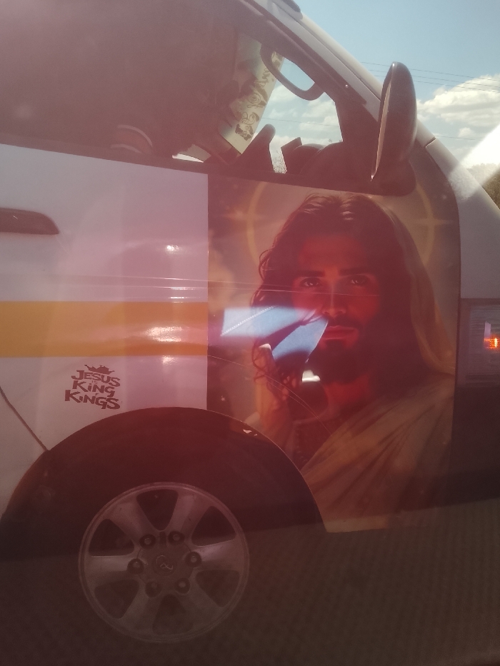 A white passenger vehicle, likely a matatu, is depicted outdoors in Kiambu Township ward, Kenya, under a clear blue sky with scattered clouds. The vehicle's side features a prominent horizontal yellow stripe. A large decal of Jesus Christ, with long brown hair, a beard, and a light-colored robe, is affixed to the vehicle's upper side, extending across a window. A faint halo or starry pattern is visible behind the depicted figure's head. A significant blue, rectangular specular reflection is present near the figure's face, possibly from a screen or reflective object. Below the yellow stripe, a smaller reddish-brown decal reads "JESUS KING KINGS" accompanied by a crown icon. A silver, multi-spoke wheel hubcap is visible at the lower left of the frame. An auxiliary rearview mirror protrudes from the upper right side of the vehicle's cab. The image exhibits a pervasive reddish-pink lens flare or reflection, particularly across the upper and central portions. No individuals or direct actions are discernible within the frame.