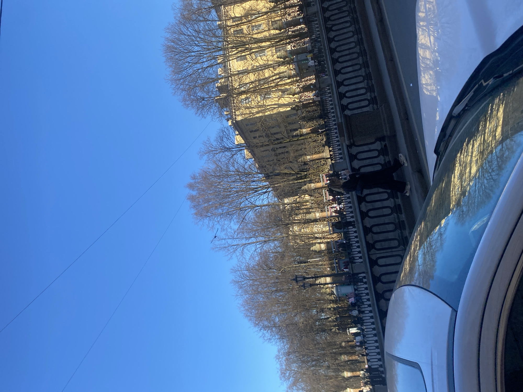 The image shows a street in Saint Petersburg, Russia, lined with bare trees and a building with many windows. People are walking along the sidewalk. A car is parked on the street. The sky is clear blue. The image is taken from a low angle, possibly from inside the car.