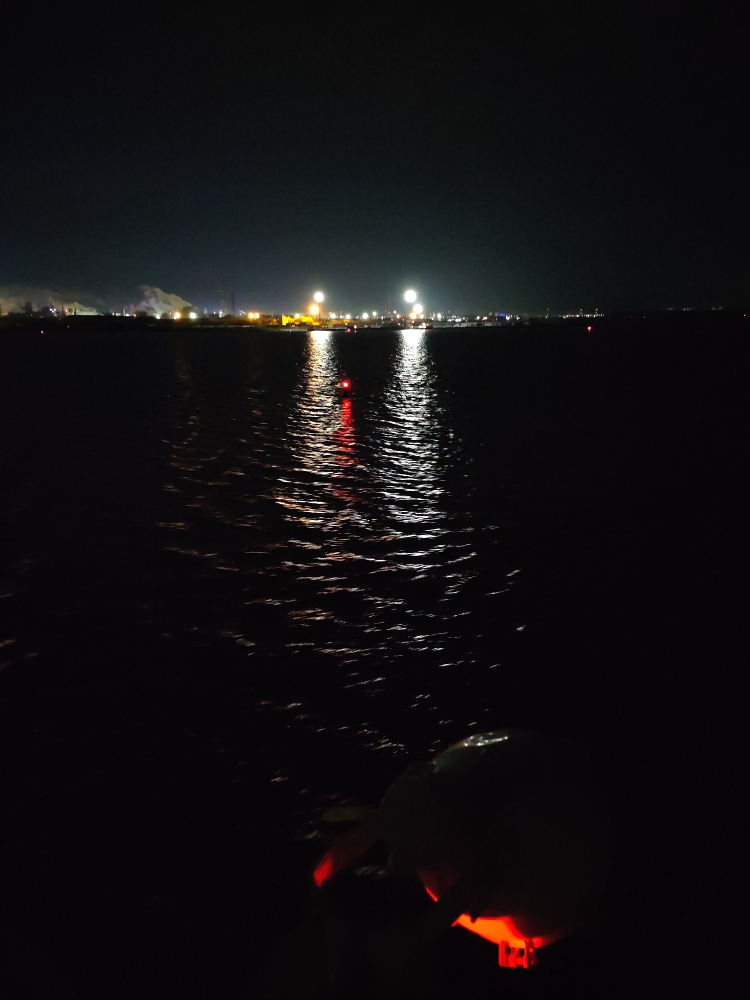 At night, the city of Unknown, Unknown presents a dark and watery scene. The horizon line is punctuated by a series of lights, mostly yellow, that appear to be city lights and perhaps an industrial complex with smoke. A red light appears in the water, and the surface of the water is rippled. The foreground is dark, with an object showing red light on it.