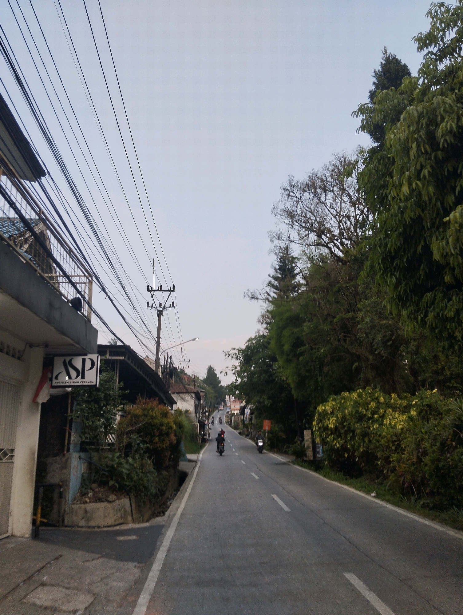 The image captures a street scene in Megamendung, Indonesia, with a view looking down a paved road that curves to the right. The sky above is a pale, hazy blue, suggesting it might be early morning or late afternoon.
On the left side of the road, a building with a white facade and a gray balcony is visible, adorned with numerous electrical wires crisscrossing overhead. A sign with "ASP" in bold lettering and smaller text below is mounted on the building. Further down the street, more buildings and vegetation line the road.
The right side of the road is densely lined with trees and lush green bushes. Several individuals are seen riding motorcycles along the road. The road itself is asphalted with white lane markings. The overall impression is that of a residential or commercial area situated on a slightly elevated terrain, characteristic of the Megamendung region.