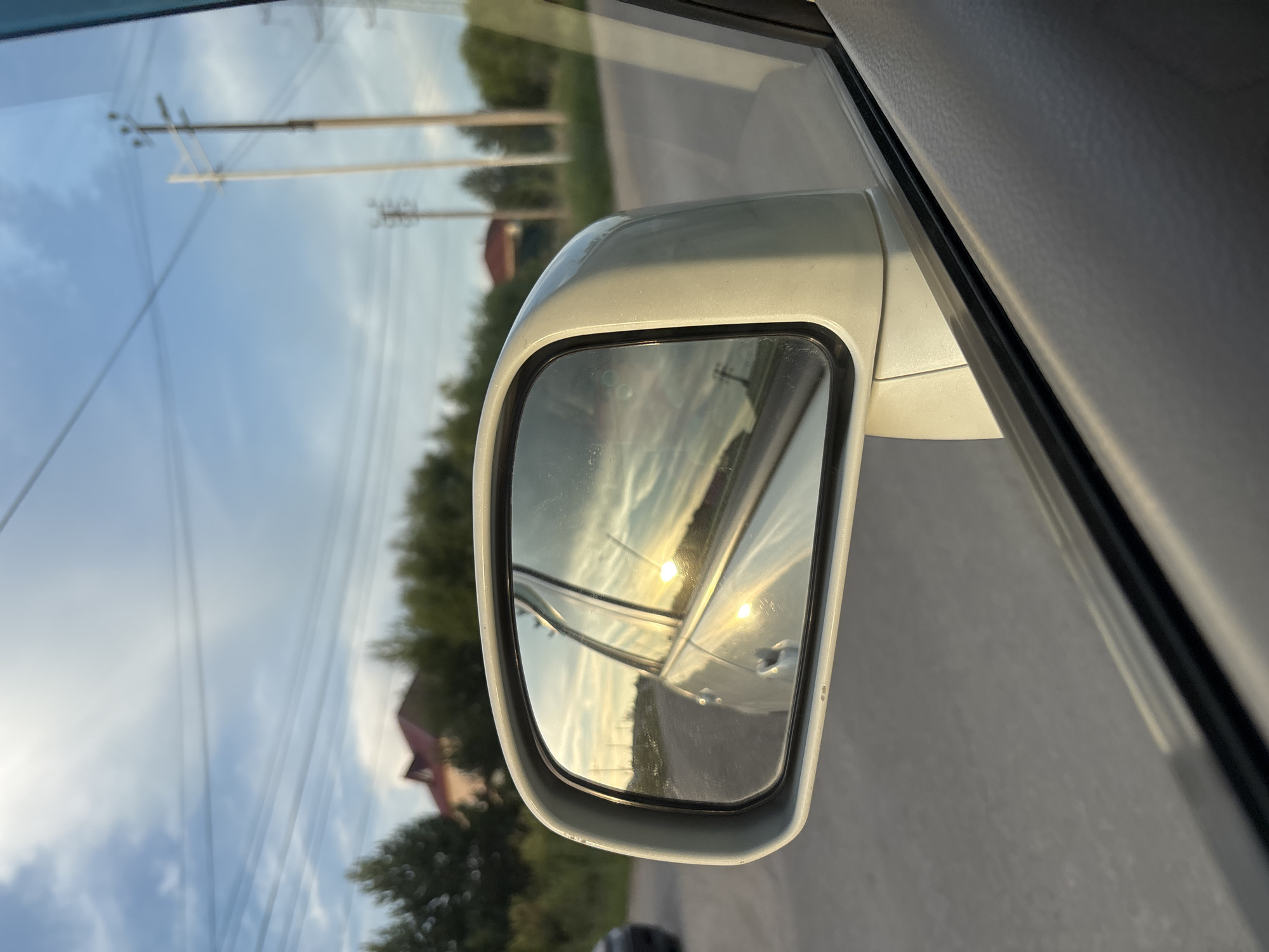 The image captures a view from inside a vehicle looking towards the side mirror. The side mirror reflects a golden sunset with clouds, and the side of the car. The exterior environment shows a road with trees and houses in the background. There are also utility poles with power lines visible. The overall scene suggests a drive through a semi-urban or rural area, possibly during the evening. The location of Chigiri, Russia is not determinable from the image itself.
