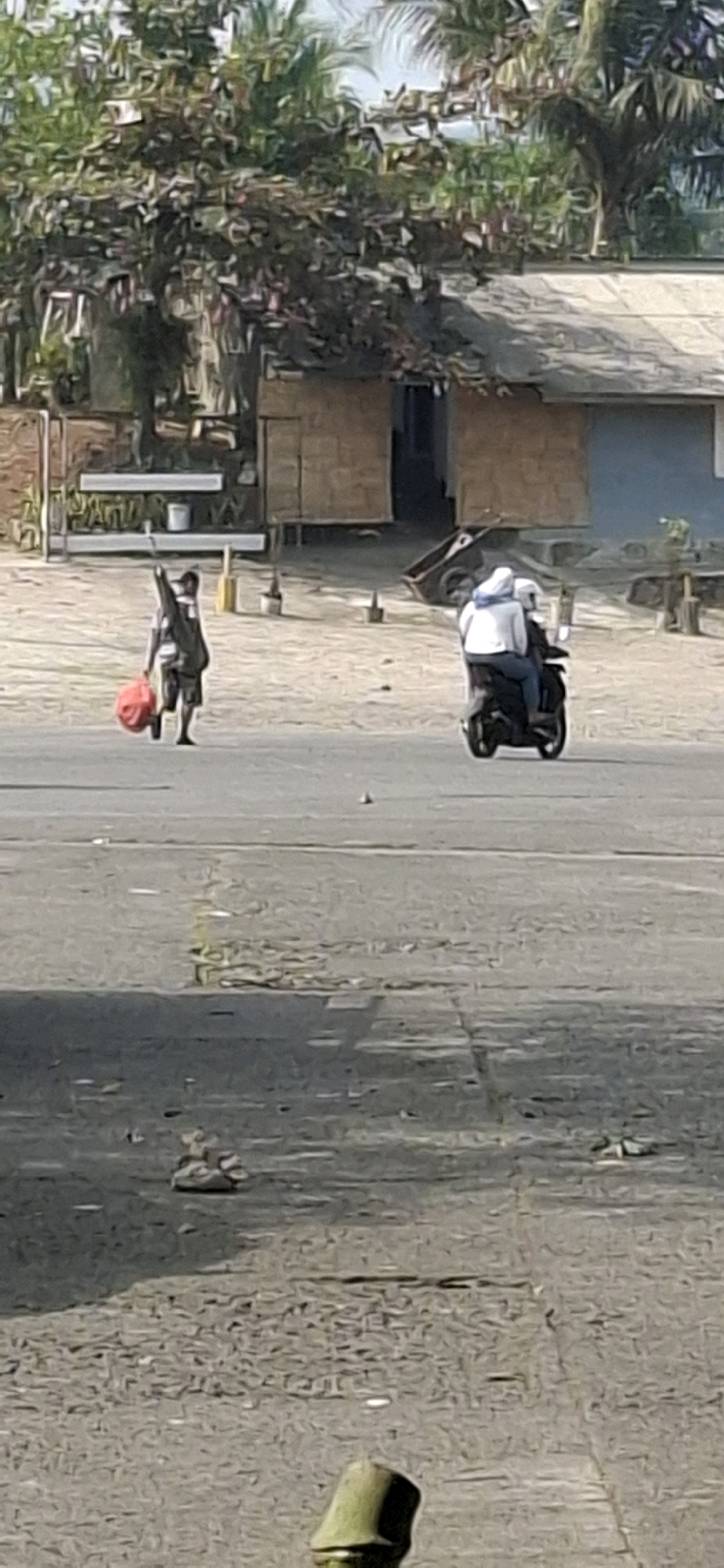 In Sirnagalih, Indonesia, on a paved surface, a person walks from left to right, carrying a large red bag in their left hand and a dark, elongated object over their right shoulder. This individual wears a light-colored shirt and dark shorts. To the right of this person, two individuals are riding a dark motorcycle, also moving from left to right. The front rider wears a white jacket and a blue head covering, while the rear rider is partially obscured. In the background, a building with a thatched roof and brick walls is visible, surrounded by trees and palm trees. The ground is a mix of pavement and sandy soil, with scattered debris.