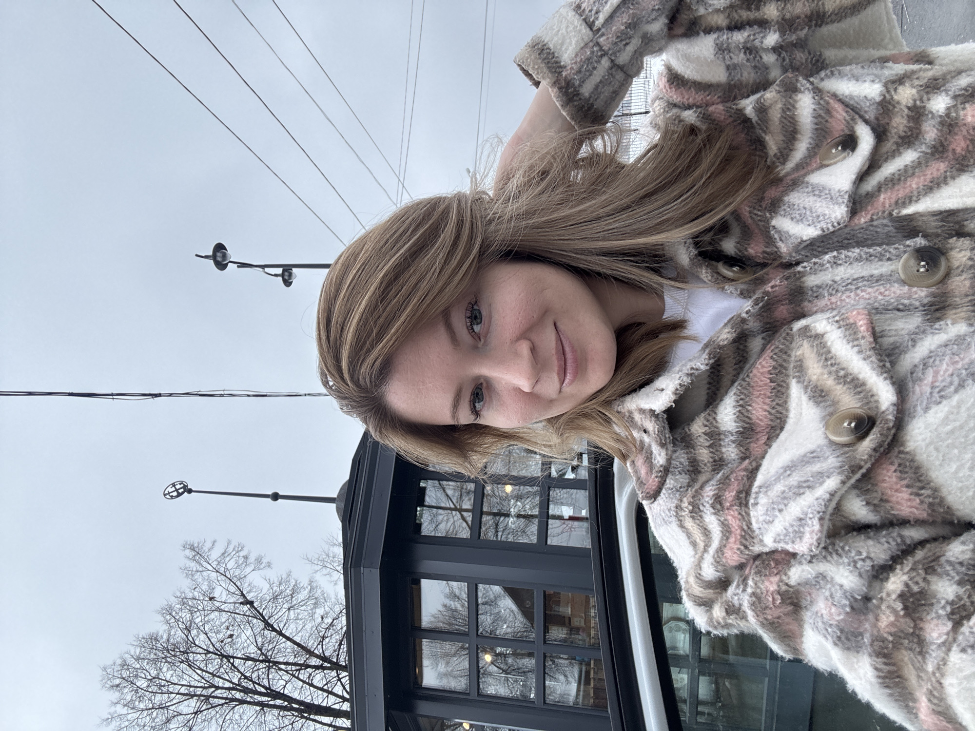 A selfie shot presents a woman with light-brown hair wearing a cream, pink, and brown plaid jacket. She's smiling slightly. The background shows a building with several windows that reflect bare trees, implying it is possibly winter or late autumn. Also in the background are utility poles and overhead wires against a light gray sky. Without specific identifying landmarks, it is impossible to confirm the exact location in Votkinsk, Russia, but the scene suggests an urban or suburban setting within the city.