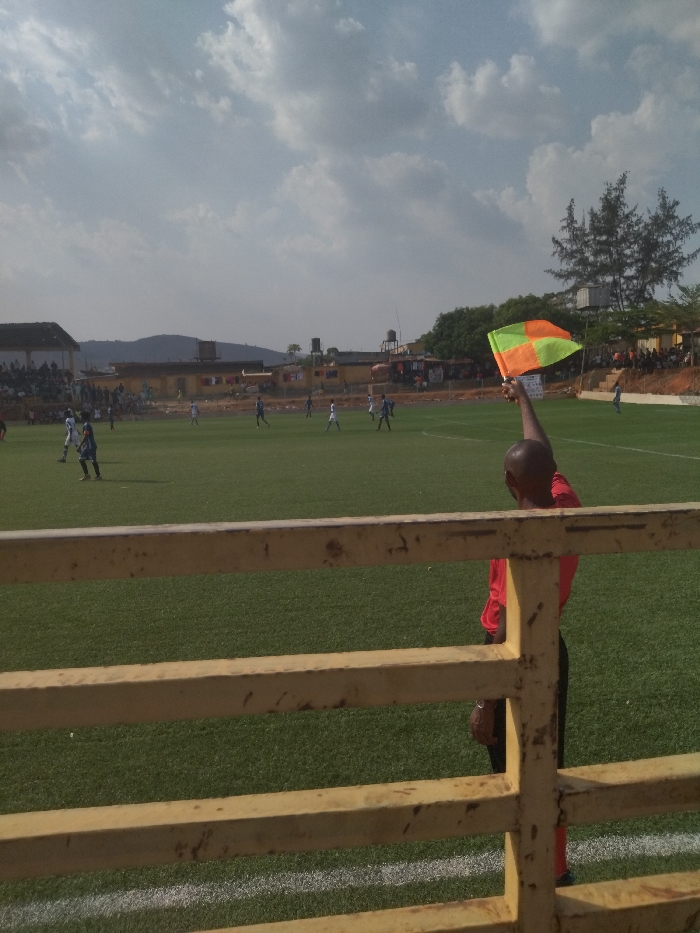 The image was taken at a soccer field in New Karu, Nigeria (latitude: 9.027, longitude: 7.578). A referee in a red shirt stands at the sidelines, raising a green and orange flag. The game is in progress. Several players are visible on the field, wearing blue and white uniforms. The spectators are sitting in the stands behind a wooden fence. The sky is partly cloudy. The field is surrounded by a fence. The image shows a match between two soccer teams. The referee is signalling something, possibly an off-side or a foul. The players are in mid-game, running and kicking the ball. The spectators are cheering and watching the game.