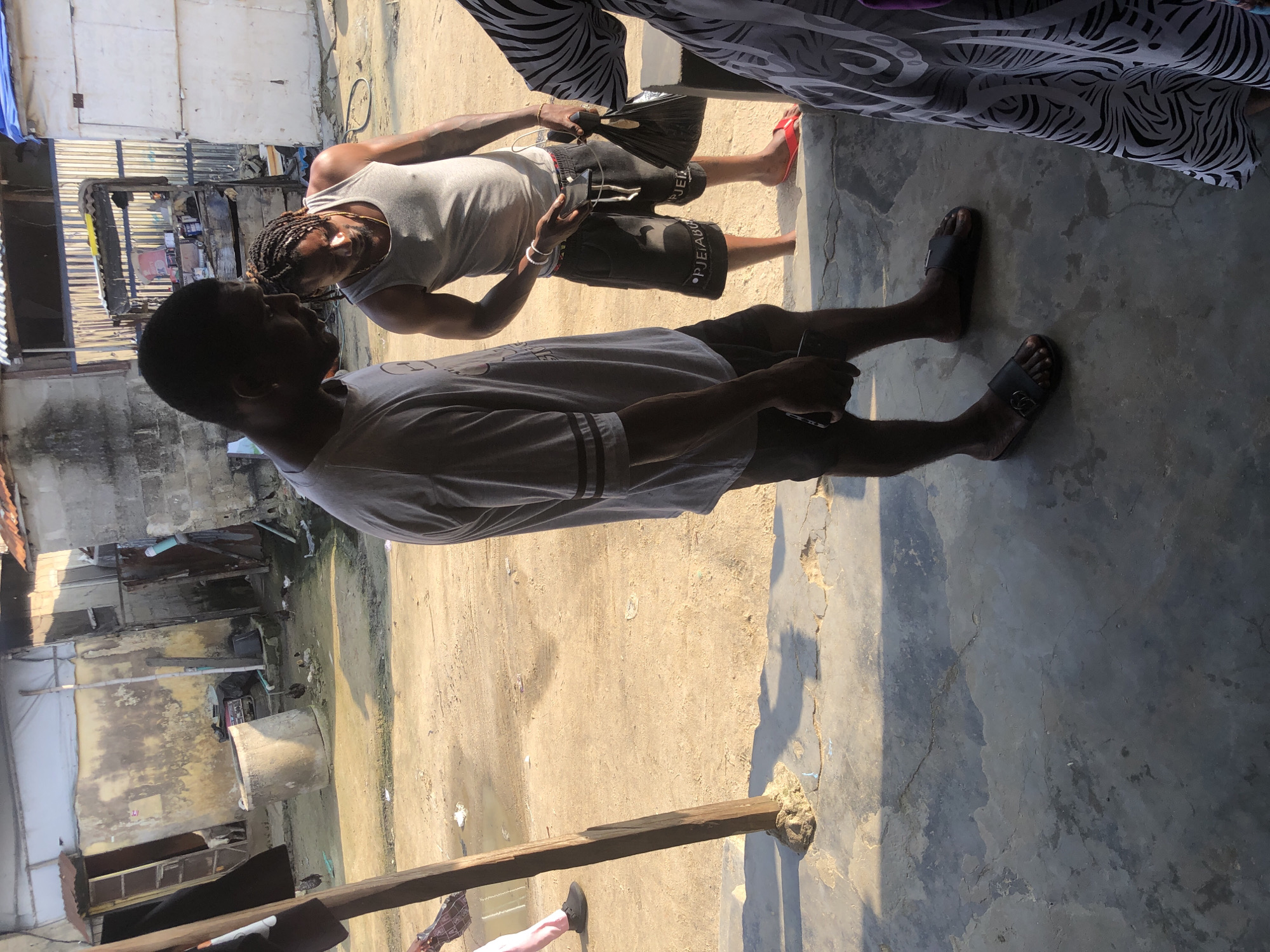 In Ado, Nigeria, two men stand near a concrete structure. The man on the left wears a gray t-shirt and dark shorts, while the man on the right wears a gray tank top and black shorts with red trim. He also has braided hair and jewelry on his wrist. The ground is dirt and concrete, with some puddles of water. In the background, there are several buildings with corrugated metal roofs and peeling paint. There is also a large concrete well and some debris scattered around. A third person, partially visible on the right, wears a black and white patterned garment.