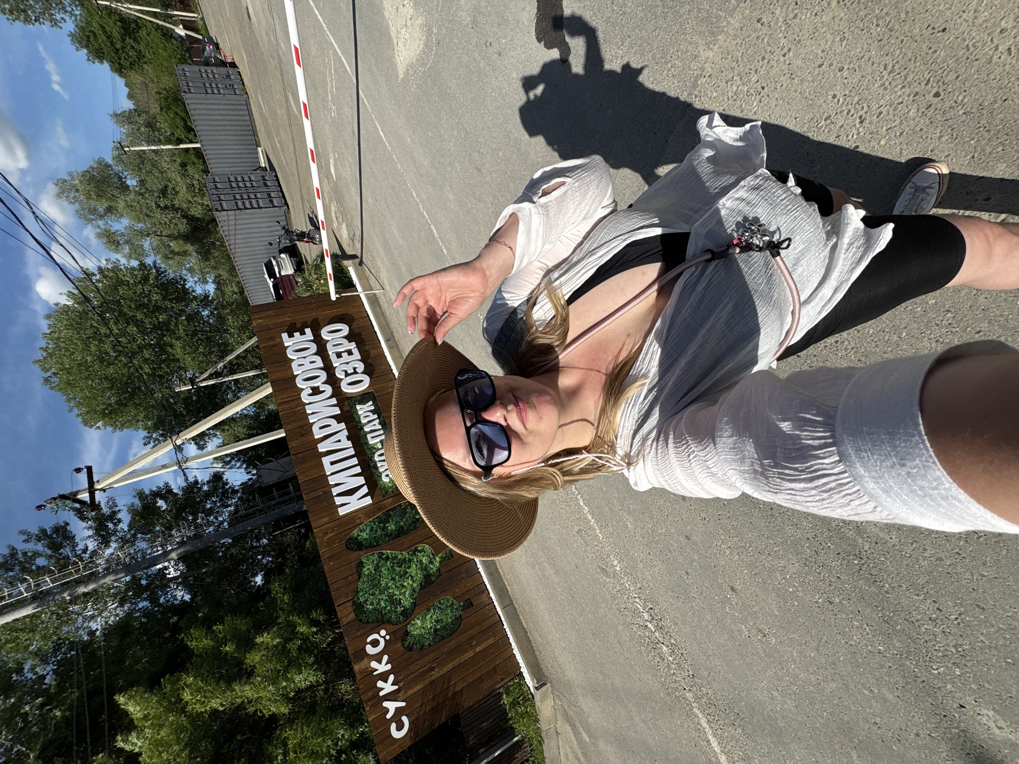 A selfie-style photo captures a woman outdoors on a sunny day, with the image rotated on its side. The woman, positioned centrally, is wearing a straw hat, dark sunglasses, a loose white sheer cover-up over a black top, black shorts, and white sneakers. A pink strap and a leash with multiple clips are visible across her body. Her expression is neutral, partially obscured by the sunglasses.
The setting is an outdoor paved area, likely a road or path. To her left, there's a prominent wooden sign with white lettering. The text clearly reads "КИПАРИСОВОЕ ОЗЕРО" (Cypress Lake), "ЭКО-ПАРК" (Eco-Park), and "СУККО" (Sukko). The sign also features green cut-outs resembling trees or foliage.
In the background, lush green trees and utility poles with power lines are visible under a blue sky with some clouds. Further back, a white and red barrier arm is present, indicating an entry or exit point. Behind the barrier, dark gray shipping containers are stacked, and a white car and a motorcycle are parked. Strong, short shadows are cast on the paved ground to the right, including the shadow of the person taking the photo, confirming bright mid-day sunlight. The scene suggests a leisurely visit to an eco-park or natural attraction.