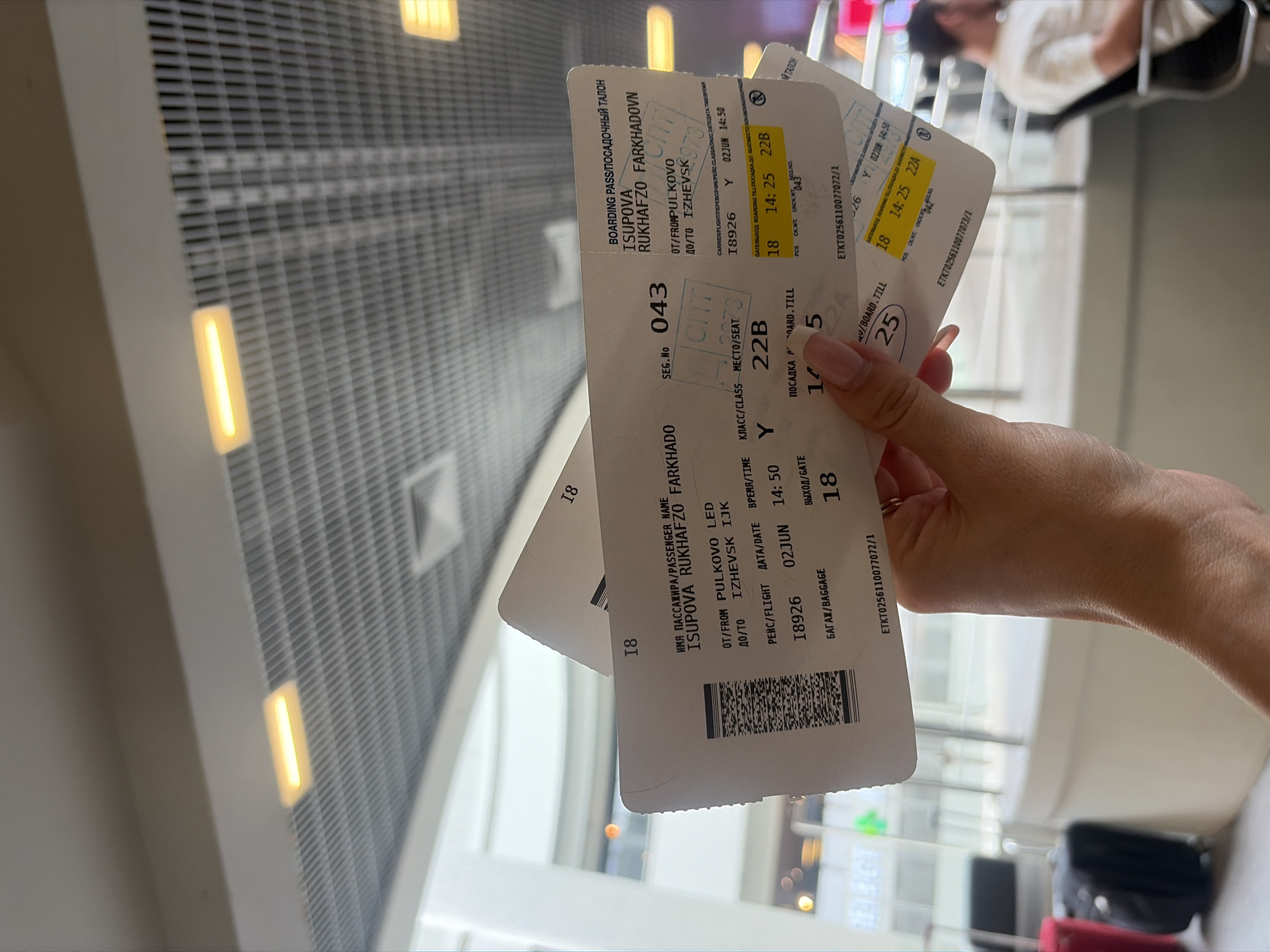 The image captures a close-up view of a person's hand holding several airline boarding passes in what appears to be an airport terminal. The hand, with light-colored manicured fingernails, prominently displays the passes, which are the central focus of the shot.
The boarding passes contain various details. One clearly visible pass shows the passenger name "ISUPOVA RUKHAFZO FARKHADOVN" and indicates a flight "OT/FROM PULKOVO LED" (Pulkovo Airport, Saint Petersburg) "ДО/ТО IZHEVSK IJK" (Izhevsk Airport). The flight number is "18926", scheduled for "02JUN" (June 2nd) at "14:50". The seat number is "22B" in "Y" (Economy) class, with a boarding gate/time indicated as "18" and "14:25". Another pass shows a seat "22A", suggesting multiple passengers or different segments. Ticket numbers "ETKT0256110077072/1" and "ETKT0256110077073/1" are also visible. Other text includes "BOARDING PASS/ПОСАДОЧНЫЙ ТАЛОН", "SEG.NO 043", "БАГАЖ/BAGGAGE", and "ВЫХОД/GATE".
In the background, the modern architecture of the airport ceiling is visible, featuring a grid-like pattern with rectangular, bright yellow light fixtures. The ambient lighting suggests it is daytime. A blurred figure of another person is partially visible sitting in the distance, and parts of what appear to be black and red luggage can be seen at the bottom right corner of the frame. The overall scene depicts a traveler holding their boarding documents while waiting in an airport.