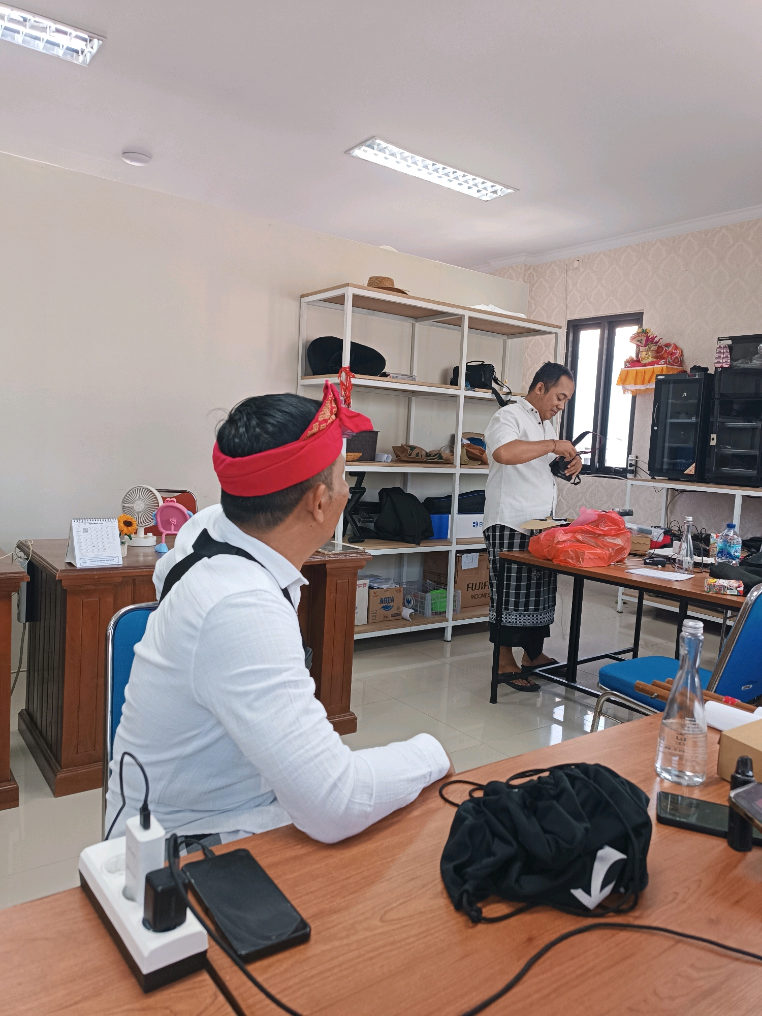 In Tabanan, Indonesia, two men are in a room. One man, wearing a white shirt and a red headband, is seated at a desk facing away from the camera. On the desk, a power strip with a white adapter and a black adapter is connected to a smartphone. Another man, in a white shirt and a black and white checkered sarong, stands at a table, handling a black object, possibly a camera. The room features white shelving units stocked with various items, including bags and boxes. A window is visible in the background, and fluorescent lights illuminate the ceiling. The environment appears to be an office or a training space.