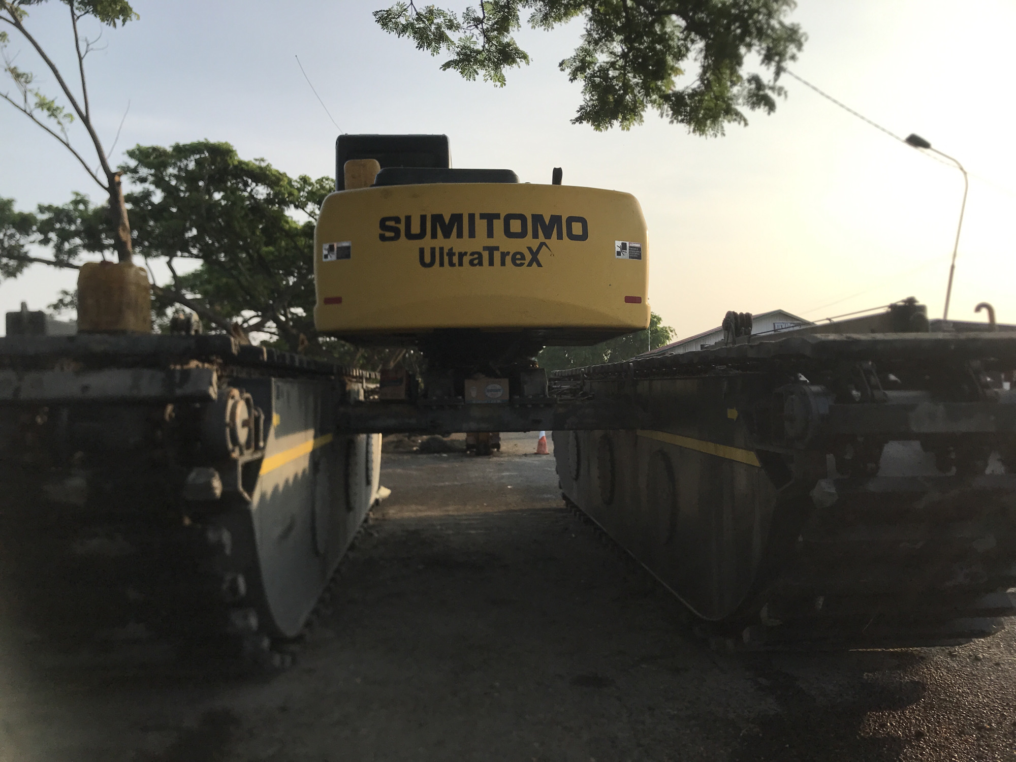 The image shows the rear of a Sumitomo UltraTreX amphibious excavator with pontoon tracks, positioned on a paved surface. The excavator is yellow with black lettering. The tracks are primarily dark in color with yellow accent stripes. The setting appears to be outdoors, possibly a construction site or an area undergoing work. Trees are visible in the background, and a street light is present on the right side. The location is reported as Lhokseumawe, Indonesia. A traffic cone is faintly visible between the pontoon tracks.
