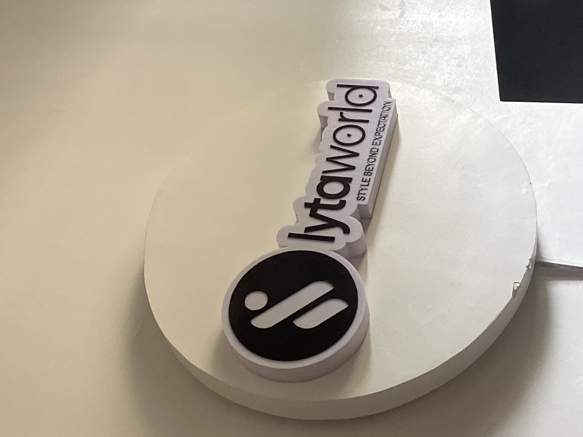 A close-up, slightly angled shot shows a sign for "lytaworld" mounted on a wall. The sign features a circular logo with a stylized "i" shape in black and white, positioned to the left of the brand name "lytaworld" in 3D purple lettering. Below "lytaworld," a smaller line of text reads "STYLE BEYOND EXPECTATION" in purple. The sign is set against a plain, light-colored wall. The bottom right of the image shows a portion of a black object, possibly a screen or monitor. The image appears to have been taken indoors. There are no people visible in the image.