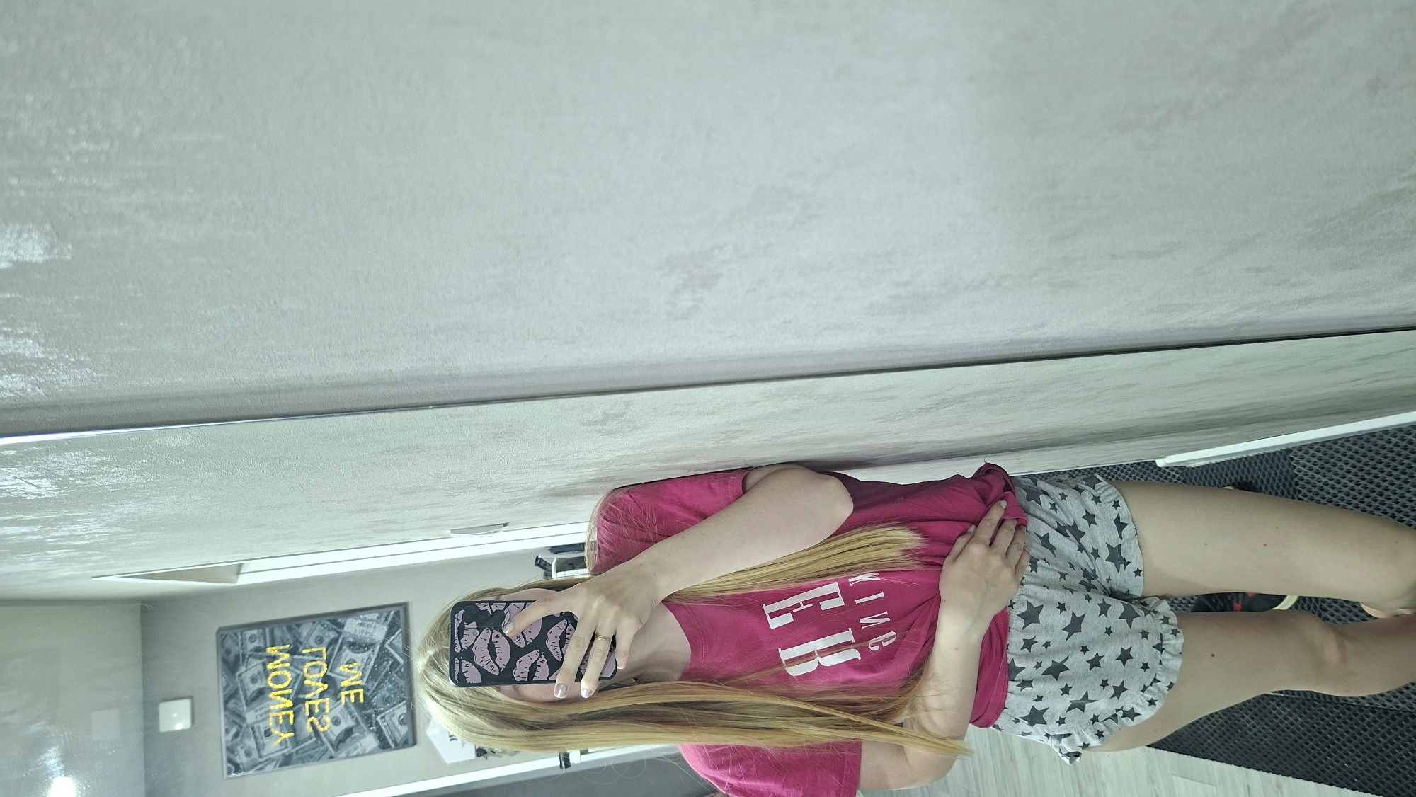 A blonde female stands indoors, likely in Chaikovsky, Russia, holding a smartphone with a lip design case in front of her face. She wears a pink t-shirt and grey shorts with black stars. A framed picture with the text "We Take Money" is visible behind her. The background shows a gray wall or surface, potentially a mirror, and a patterned floor mat. The image appears to be a mirror selfie.