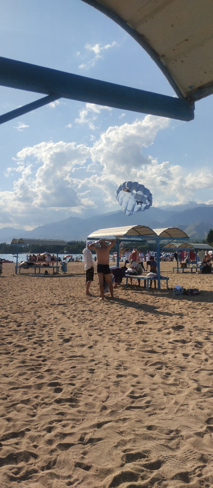 This is a daytime, eye-level, medium shot of a sandy beach with a body of water and mountains in the background. The sky is blue with large, fluffy white clouds. A blue and white parachute is visible in the sky, suggesting a parasailing activity. In the foreground, the sand is disturbed with footprints. In the middle ground, there are several figures, including a man in black swim trunks with his hands on his head, another man in a white shirt and dark shorts, and other people sitting or standing under shaded structures. There are benches and tables set up on the beach. The water appears calm, and beyond it, trees line the shore before a range of mountains. The overall scene depicts a recreational area, likely a resort, on a sunny day. The location is identified as Cholpon-Ata, Kyrgyzstan, a resort town on the northern shore of Issyk-Kul Lake.