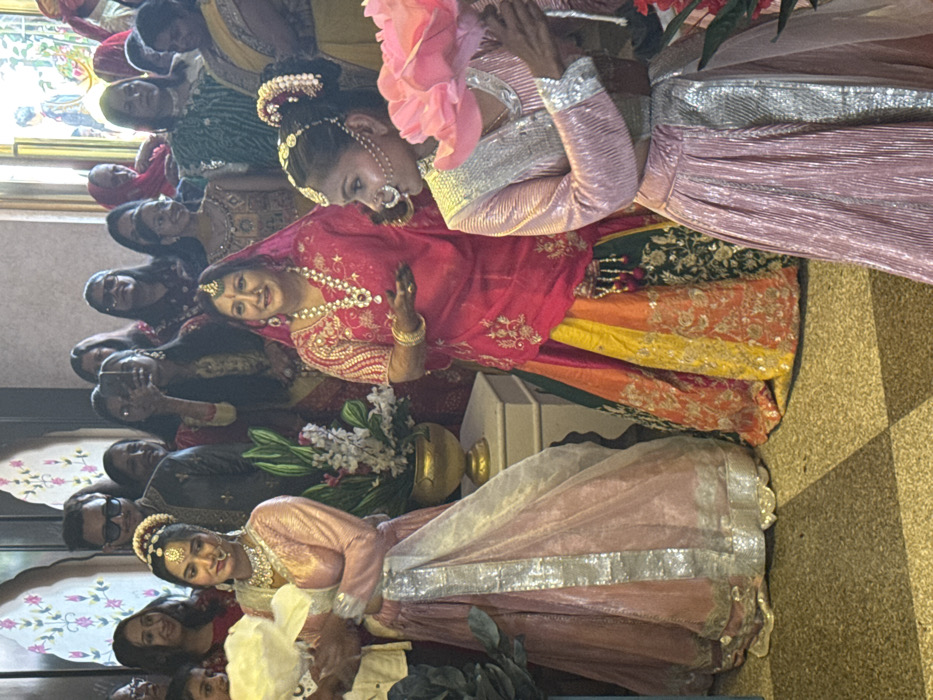 A group of people, predominantly women, are gathered. Several individuals wear traditional Indian attire. One woman in a bright pink top and silver skirt stands out. Beside her, another woman wears a red top with a multi-colored skirt of yellow, orange, and green. Flowers in a gold vase rest between them. The crowd appears to be celebrating a special event.