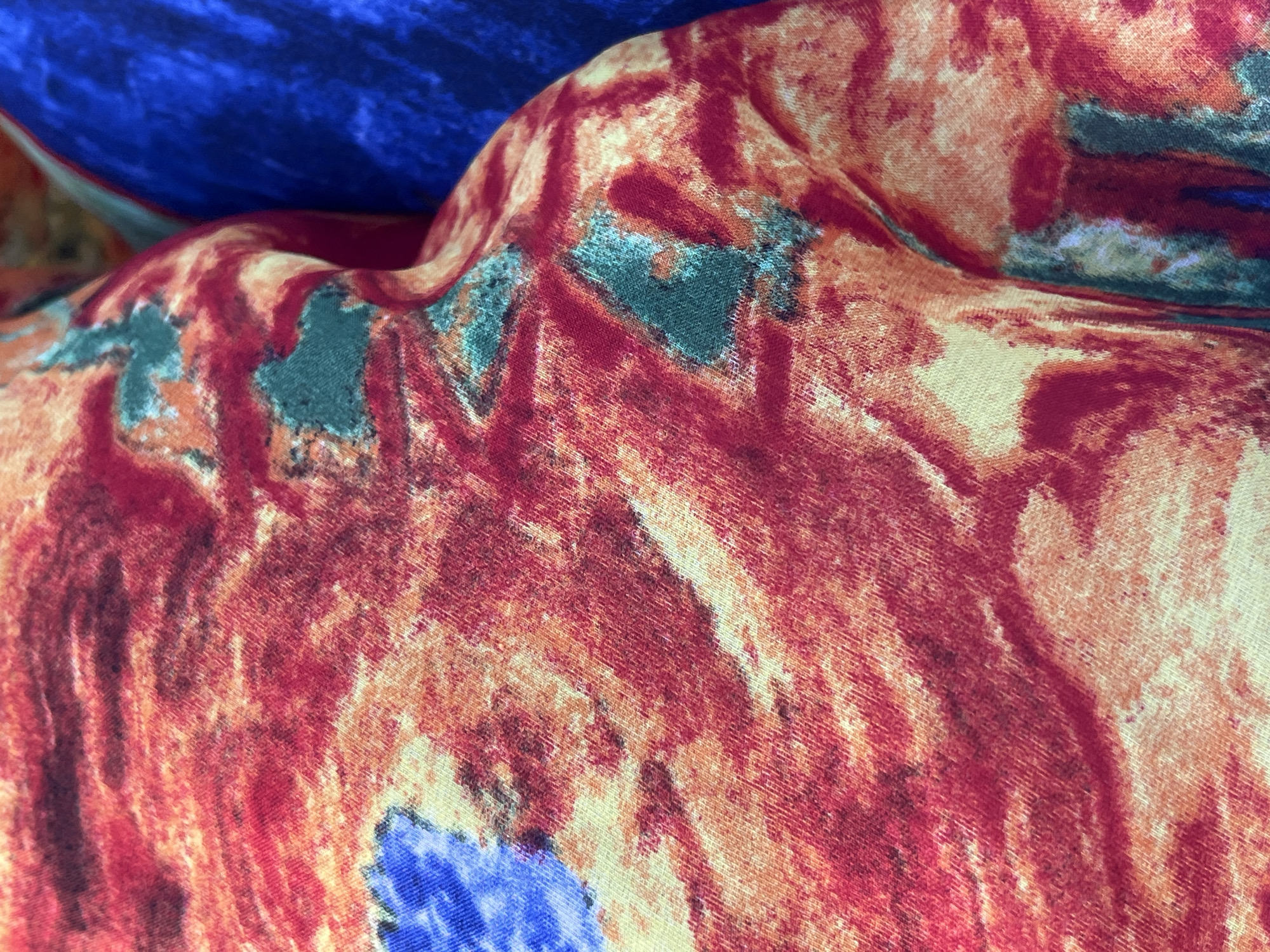 The image shows a close-up view of a textured fabric with a vibrant, abstract pattern of reds, oranges, and hints of blue and green. The fabric appears to be draped or folded, creating shadows and highlights that emphasize its texture. In the background, a section of a solid blue fabric is visible. The scene is tightly cropped, offering no context of its surroundings or location. There are no people or animals present. There is no information that can be used to determine the location in the city of Pistula, Montenegro.