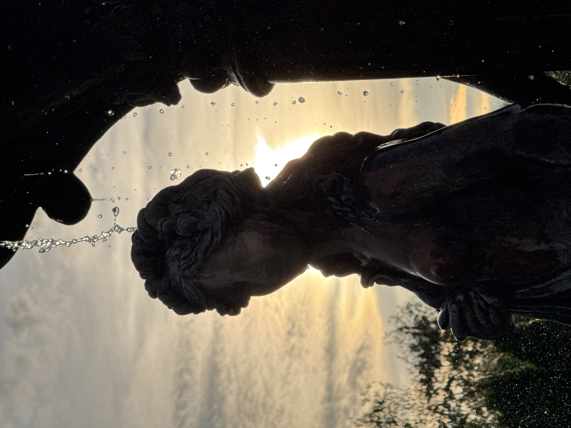 An outdoor photograph depicts a portion of a bronze fountain, silhouetted against a dramatic sunset or sunrise sky. The primary subject is a neoclassical-style female statue, rendered in dark silhouette, positioned slightly left of center. The statue's head, featuring intricately styled hair, is bowed, and her profile is partially visible. Her upper torso is exposed, suggesting nudity or partial drapery. To her right, a large, dark, ornate element of the fountain structure is visible, from which streams of water cascade downwards. Numerous individual water droplets are suspended in the air, some illuminated by the intense backlighting from the sun, which appears as a bright, starburst-like point behind the statue's head and right shoulder. The sky transitions from a bright golden-orange at the horizon to lighter greyish clouds above, displaying subtle striations. In the lower background, dark, indistinct foliage or trees are faintly discernible. The fountain depicted is the "Girl with a Swan" (also known as "Nymph with a Swan") located in Alexandrovsky Square (also known as Central Square Garden) in Minsk, Belarus.