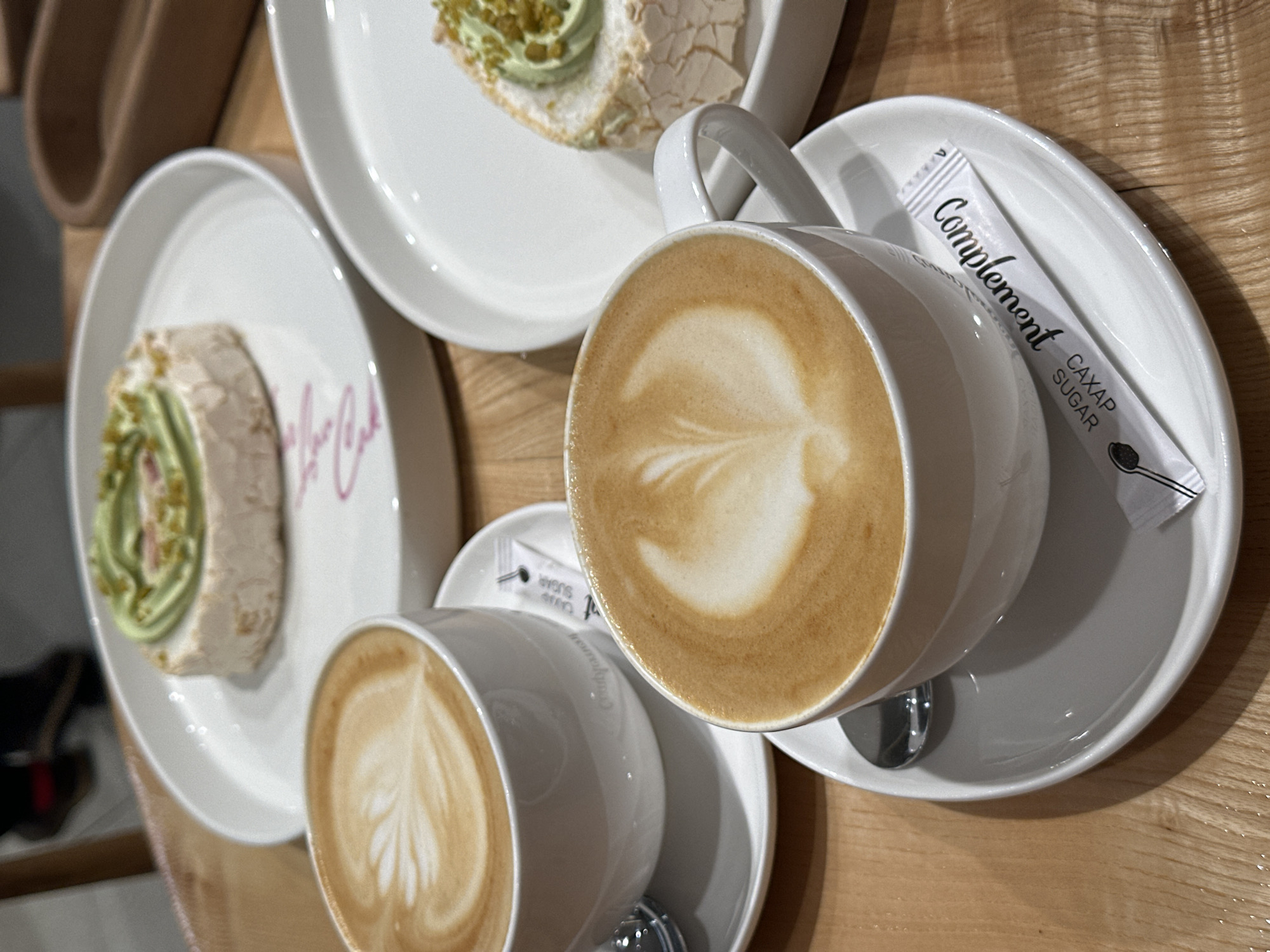 The image shows a table setting with two cups of latte and two dessert plates. The table appears to be made of light-colored wood. One plate contains a slice of a meringue roll with pistachio cream. Another dessert is a sandwich with a white base and green topping. The latte cups have latte art and sit on their respective saucers. A sugar packet labeled "Complement CAXAP SUGAR" in Cyrillic is on one saucer. The setting is located in Rostov-on-Don, Russia.