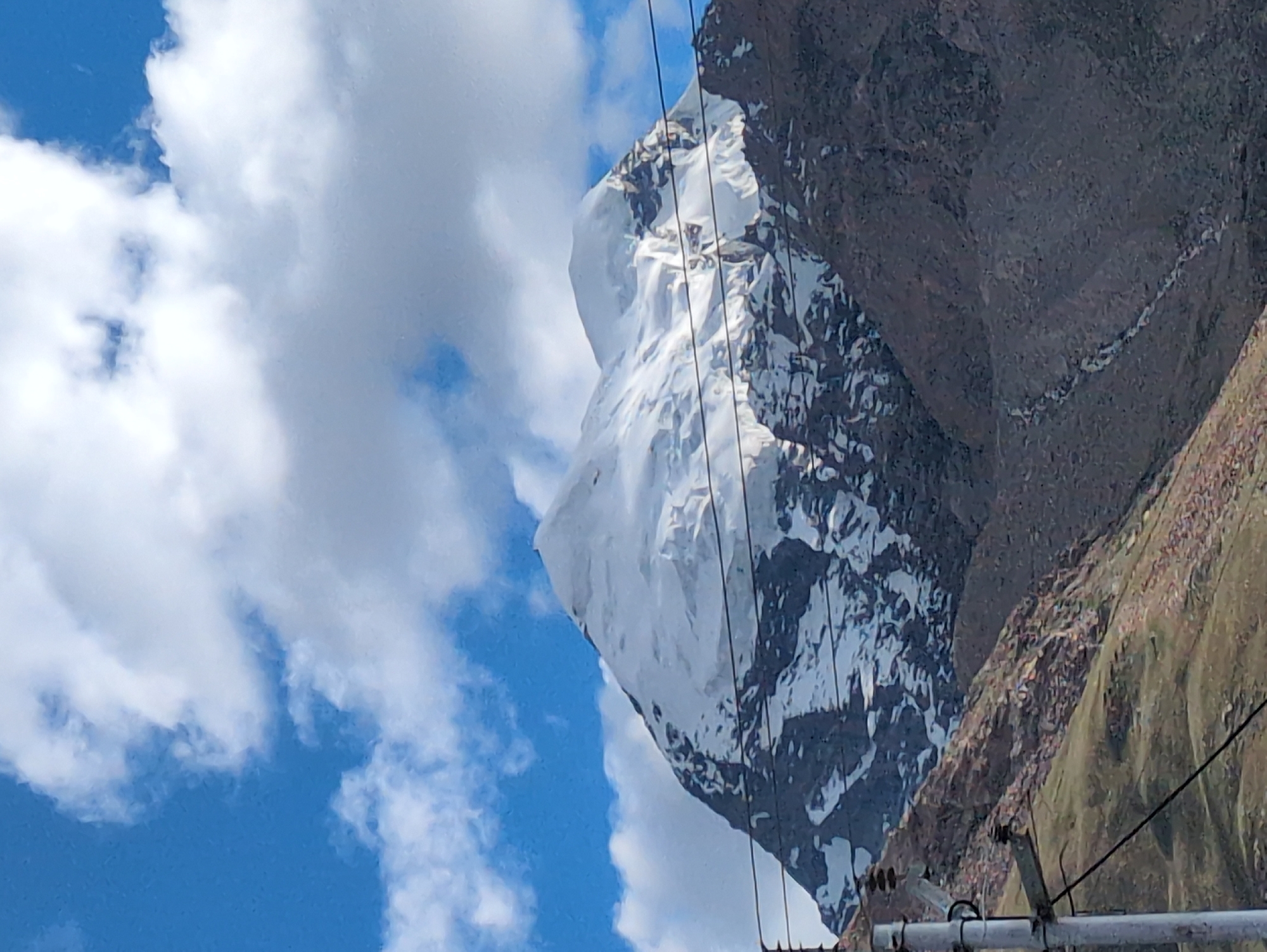 This image captures a majestic snow-capped mountain peak against a vibrant blue sky dotted with white clouds. The mountain exhibits rugged, rocky terrain with patches of snow and ice, suggesting a high-altitude environment. In the foreground, a utility pole with electrical wires is visible, indicating the presence of infrastructure near the natural landscape. The lower slopes of the mountain are covered in dry vegetation and rocky scree. The scene is likely located in or near Padum, India, a town situated in the Zanskar region of Ladakh, known for its dramatic mountainous scenery.