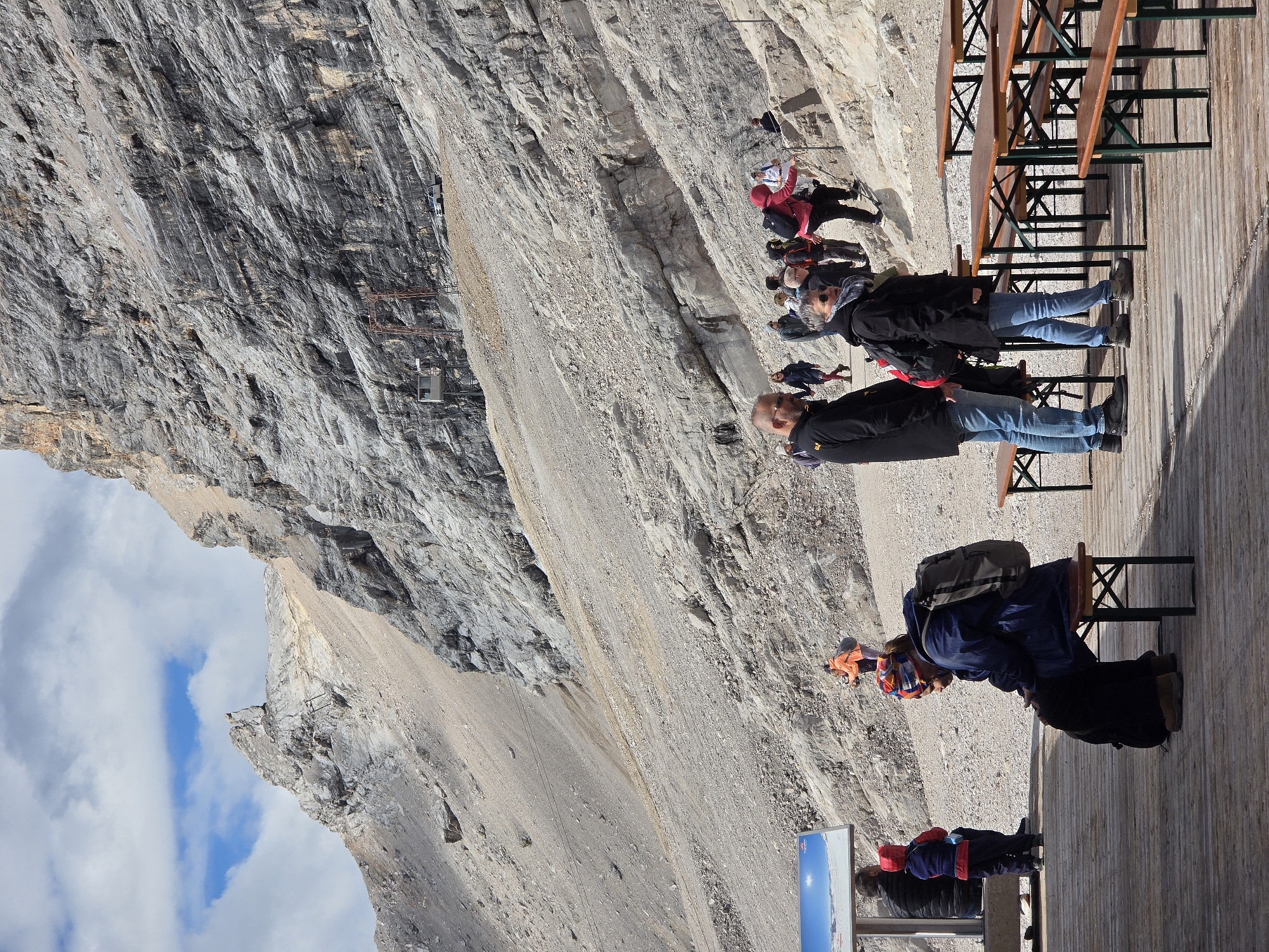 The image depicts a mountain landscape with a group of people in the foreground and midground. The background features a steep, rocky mountain face under a cloudy sky. In the foreground, several people are gathered around picnic tables on a wooden platform. Some are seated on benches, while others are standing. Further up the rocky slope, more individuals are walking or hiking, some with backpacks. The environment appears to be a tourist area at a high altitude, likely associated with a mountain resort or viewing point.
While the image itself does not directly indicate the city of Garmisch-Partenkirchen, Germany, it is highly probable that this scene is located on the Zugspitze mountain, Germany's highest peak, which is accessible from Garmisch-Partenkirchen and features similar rocky terrain and infrastructure for tourists, such as cable cars and viewing platforms. The presence of picnic tables suggests a designated rest or dining area for visitors.