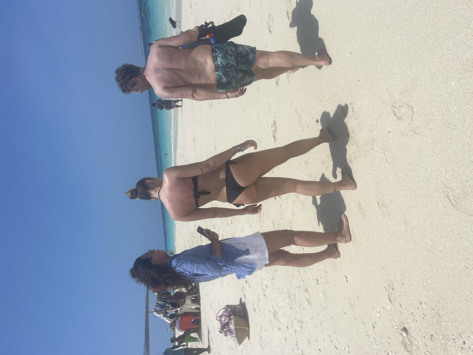 This image depicts a bright, sunny day on a white sandy beach in Zanzibar, Tanzania. Three individuals are prominently featured in the foreground, facing towards the ocean.
On the left, a woman with dark, curly hair wears a light blue long-sleeved shirt over white shorts. She stands barefoot, holding a black smartphone in her right hand, and looks towards the right side of the frame. To her right, a second woman, seen from behind, wears a black strapless bikini top and matching thong-style bottom. Her hair is gathered in a bun, and she has several bracelets on her left wrist and a small dark tattoo on her left lower calf. She stands barefoot, facing the ocean. To the right of the second woman, a man is seen from behind, shirtless, wearing dark green patterned swim shorts. He is barefoot and carries a black bag, likely containing flippers and snorkel gear, over his left shoulder as he walks towards the ocean. Distinct shadows of these individuals are cast on the sand in front of them.
In the mid-ground on the left, a makeshift shelter or boat canopy provides shade for several people seated on white and red plastic chairs. A woven basket with items is visible on the sand near the left foreground woman. The background reveals a clear, light blue sky overhead, transitioning to turquoise and then deeper blue ocean water. Distant figures and objects are visible in the water and along the far shoreline.