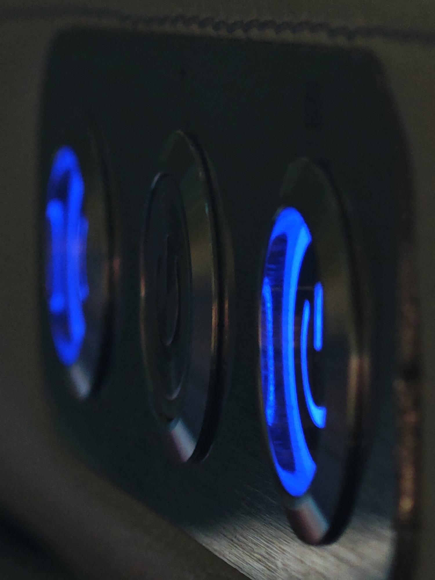 The image is a close-up, dimly lit, high-angle shot of three circular, metallic buttons on a dark surface. The buttons are illuminated from within by blue LED lights. The leftmost button has an abstract, curved blue light pattern. The middle button shows a subtle, embossed power symbol illuminated by a faint blue light. The rightmost button displays a more distinct, U-shaped blue light pattern. The surface appears to be made of brushed metal or a similar material. The image does not provide any discernible information about the city of Bờ Y, Vietnam, or any environmental or human context.