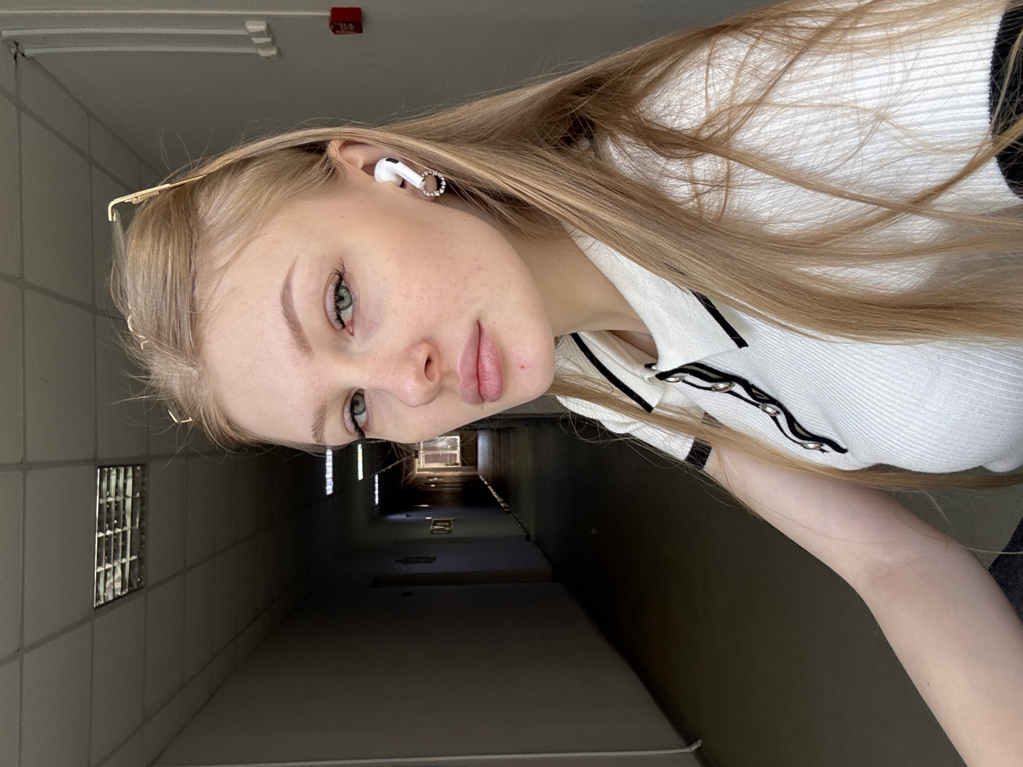 A young woman with long blonde hair and fair skin is in a hallway. She is wearing a white, collared shirt with black trim. She has an earbud in her left ear and a gold hair clip on her head. The hallway is long and dark, with square windows along the left wall and overhead lights in the distance. The woman is looking towards the camera with a slight tilt of her head. The location is in Perm, Russia.