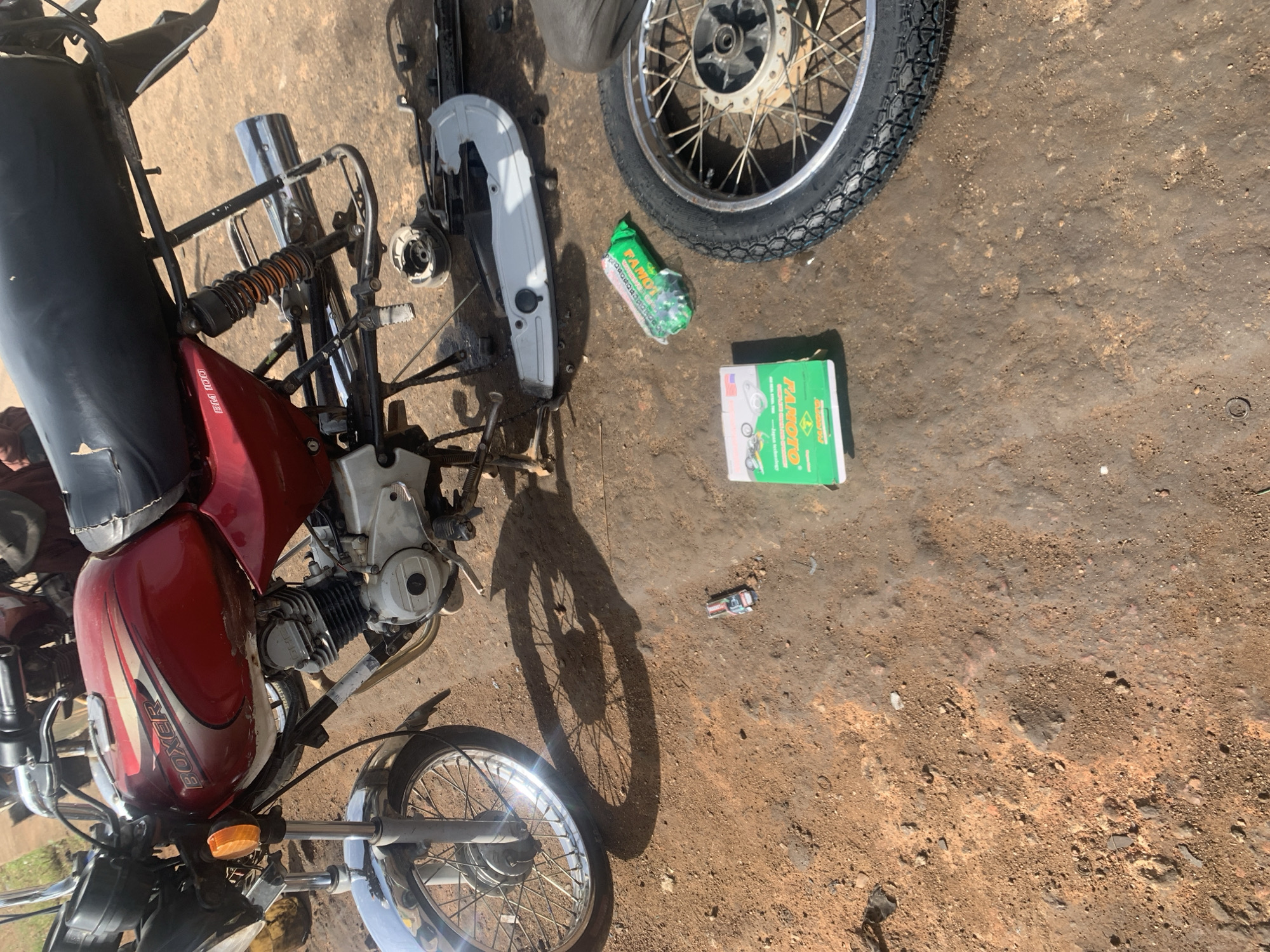 In Gura Top, Nigeria, a red and silver Boxer motorcycle is parked on a dirt and gravel surface. The motorcycle is partially disassembled, with its front wheel removed and lying on its side. A spare tire and rim are also visible, as well as a silver chain guard and other motorcycle parts. A green box and a green plastic bag are on the ground near the motorcycle. The scene appears to be a roadside repair area, with other motorcycles and people visible in the background.