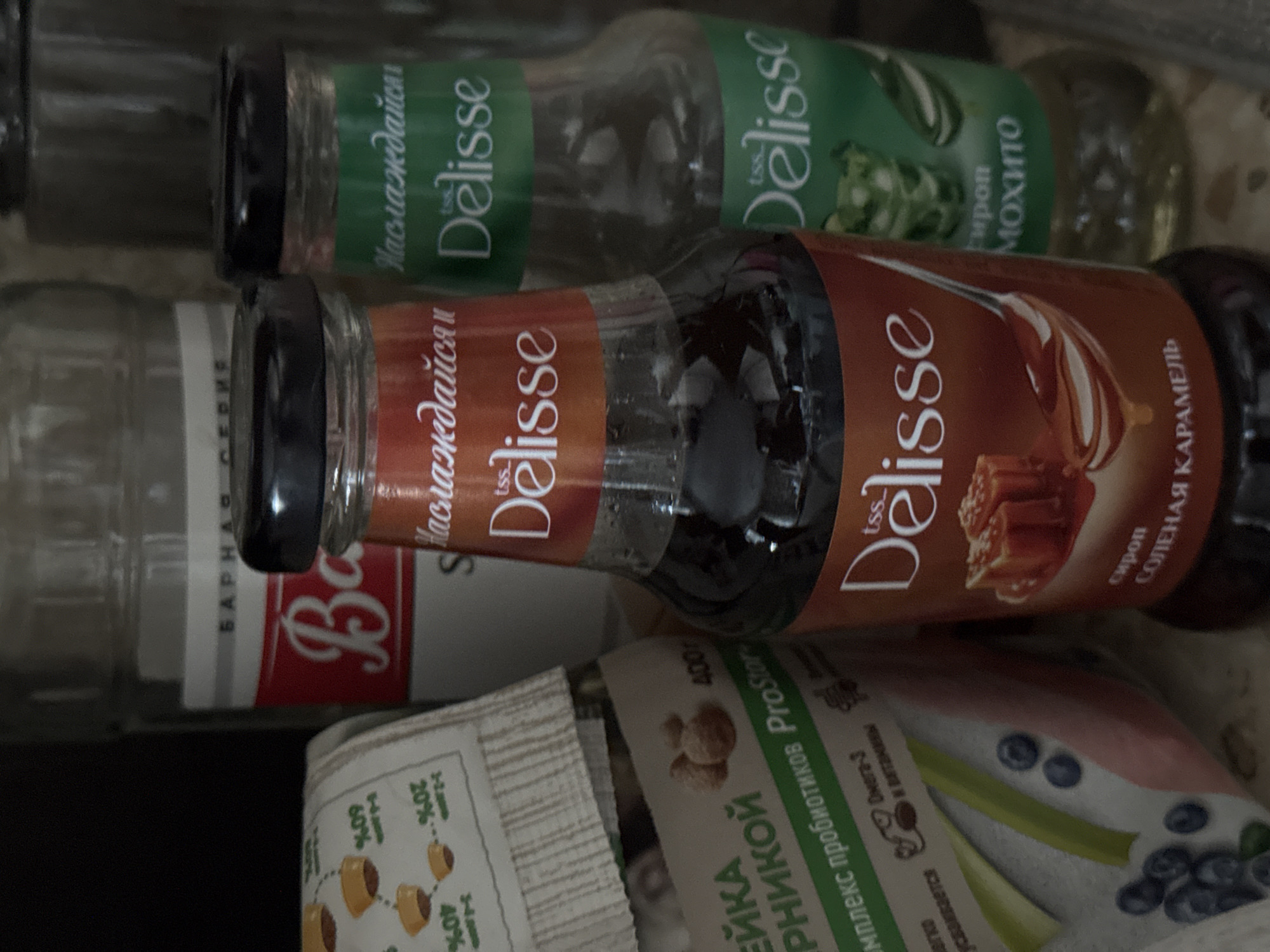 The image shows a close-up, overhead view of several bottles and packaging. In the foreground, a bottle of "Delisse" caramel syrup is prominent, with text in Russian indicating "СОЛЕНАЯ КАРАМЕЛЬ" (salted caramel). Behind it, another bottle labeled "Delisse" is visible, this one with green text suggesting "Сироп Мохито" (Mojito syrup). To the left, part of a clear glass bottle with a red and white label is visible, with the text "БАРНА СЕРИЯ" (Barna Series) and a large "Ba" in cursive. Below these bottles, there is packaging with Russian text, including "комплекс пробиотиков ProStore" (probiotic complex ProStore), and percentage markings like "20%" and "60%," along with illustrations of small, brown capsules or pills. The overall impression is of a collection of food or beverage products, possibly in a kitchen or pantry setting, given the domestic feel. The lighting is somewhat dim and diffused, suggesting indoor conditions, possibly at night or in an area with limited natural light. The items are arranged in a slightly cluttered, casual manner.