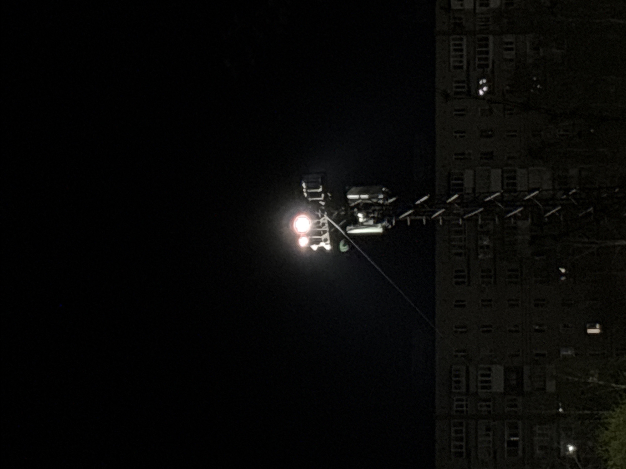 The image, taken at night, shows a building in Chaikovsky, Russia, illuminated by floodlights mounted on an extended aerial work platform. The building is a multi-story residential structure with numerous windows. The work platform is positioned close to the building, with the lights directed towards it. A cable extends from the platform. The surrounding environment appears dark.