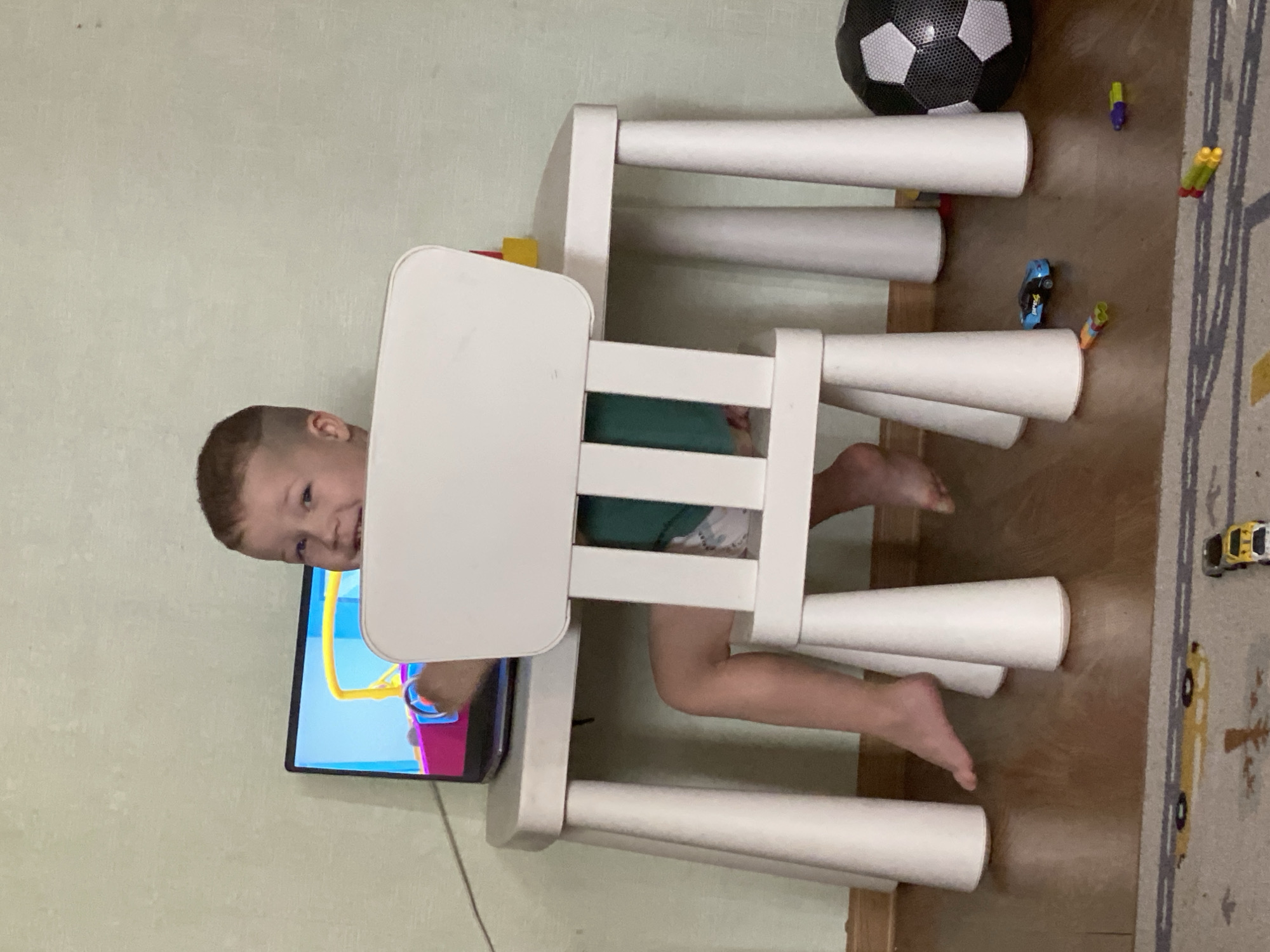 A child is stuck inside a white table/chair set. The child is wearing a green shirt and patterned shorts. The child's head is turned toward a TV screen with a cartoon playing. A soccer ball, toy cars, and toy darts are on the floor near the table set. The floor has both wood and a rug with car lanes on it. The wall is light green. The exact location in Sochi, Russia is undeterminable without more information.
