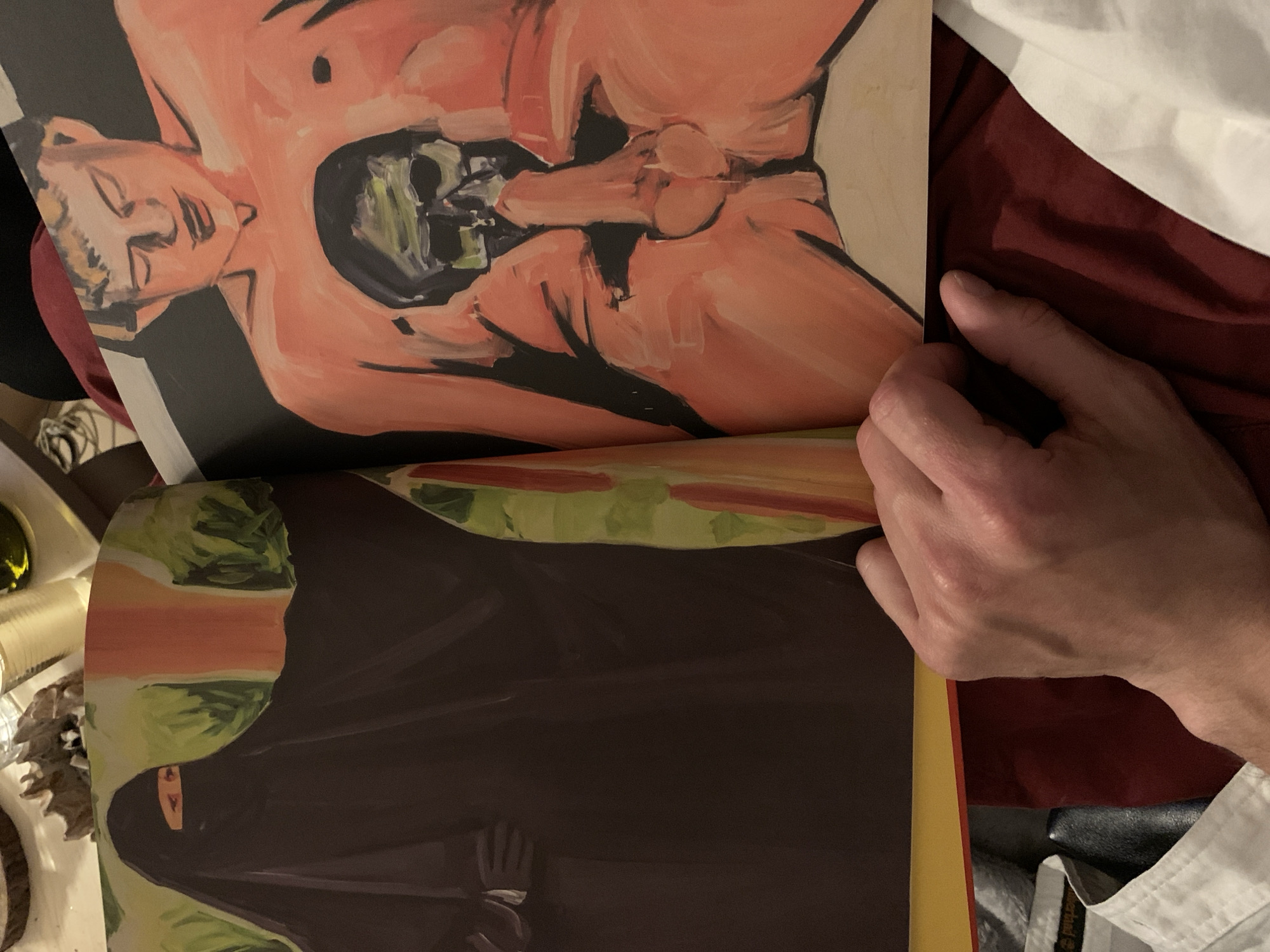 A person's hand is holding open a book or magazine with two pages visible. The top page contains a painting of a person lying down with their mouth open, and within their mouth is a depiction of a skeletal figure. The skin tone is predominantly pink, with dark shading defining the contours. The bottom page shows a painting of a figure draped in a dark, possibly black, full-body covering typical of some Islamic attires, with a small slit revealing the eyes. The background of the bottom painting includes hints of green and reddish-brown. The background around the book shows elements of a table or surface with various items scattered around. There is no indication of the location being Tel-Aviv, Israel.