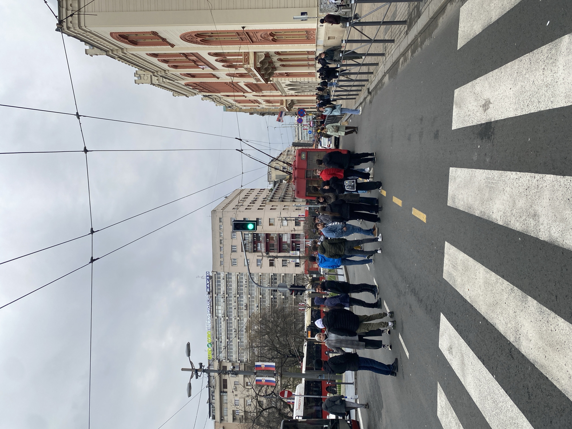 A street scene in Belgrade, Serbia, shows a group of pedestrians crossing a zebra crossing at a street intersection. A red trolleybus is near the intersection. Overhead wires for the trolleybus are visible. In the background, various buildings are present, including one with a green and white sign that includes the word "RAPID." The Serbian flag is visible on a pole near the trolleybus. The sky is overcast. A traffic light shows a green signal. People are standing along a railing on the opposite side of the street.