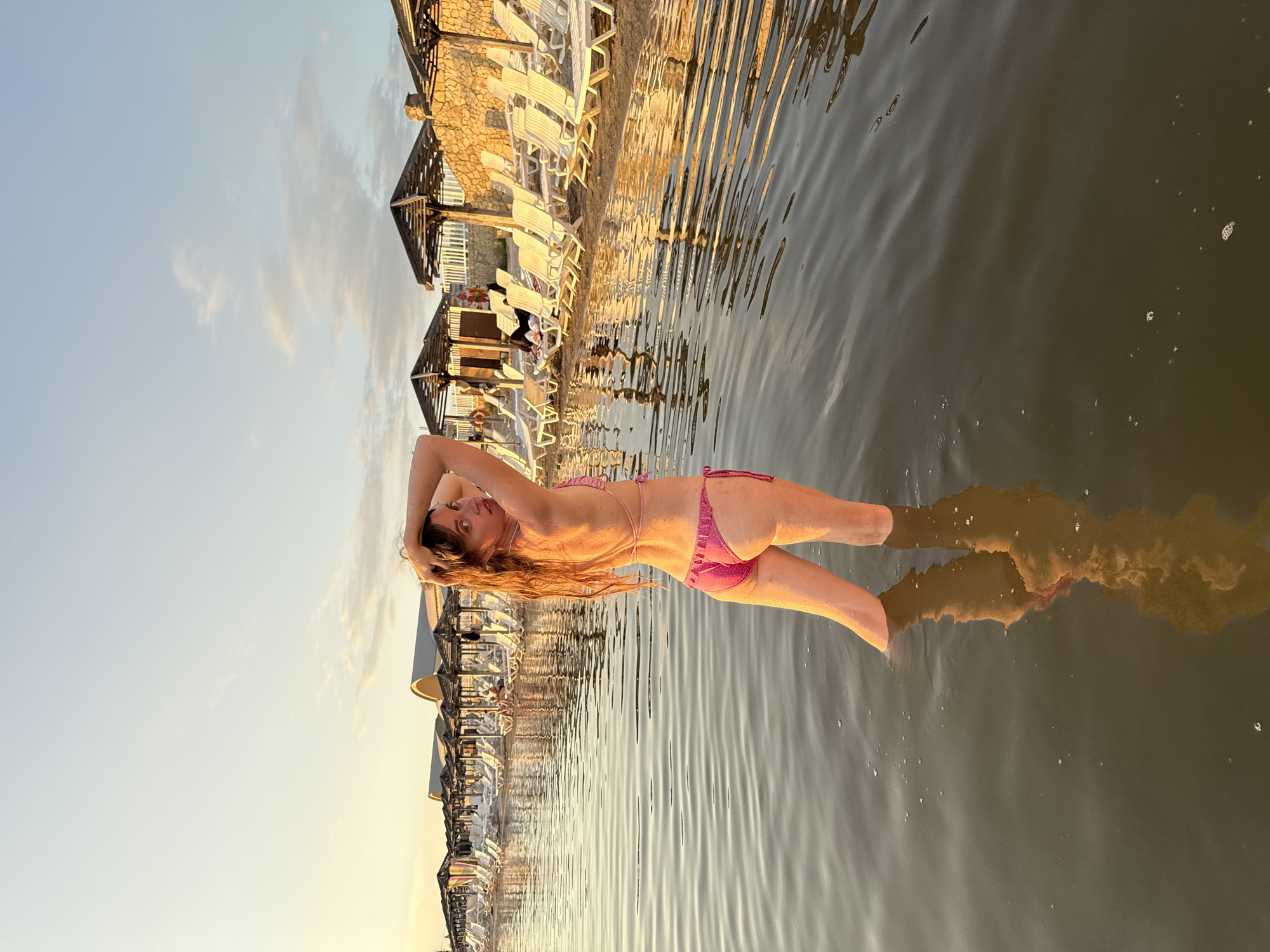 A woman stands in shallow, dark water, facing away from the camera and looking over her shoulder. She is wearing a pink, polka-dotted bikini. Her hands are behind her head, and her hair is long and reddish-brown. The water is calm, with ripples and small bubbles visible. In the background, a beach with rows of white lounge chairs and thatched umbrellas is lined with buildings. The scene appears to be at a resort or beach club in Sol-Iletsk, Russia, possibly on a salt lake given the region's reputation. The lighting suggests it is either sunrise or sunset, with a warm glow illuminating the scene.