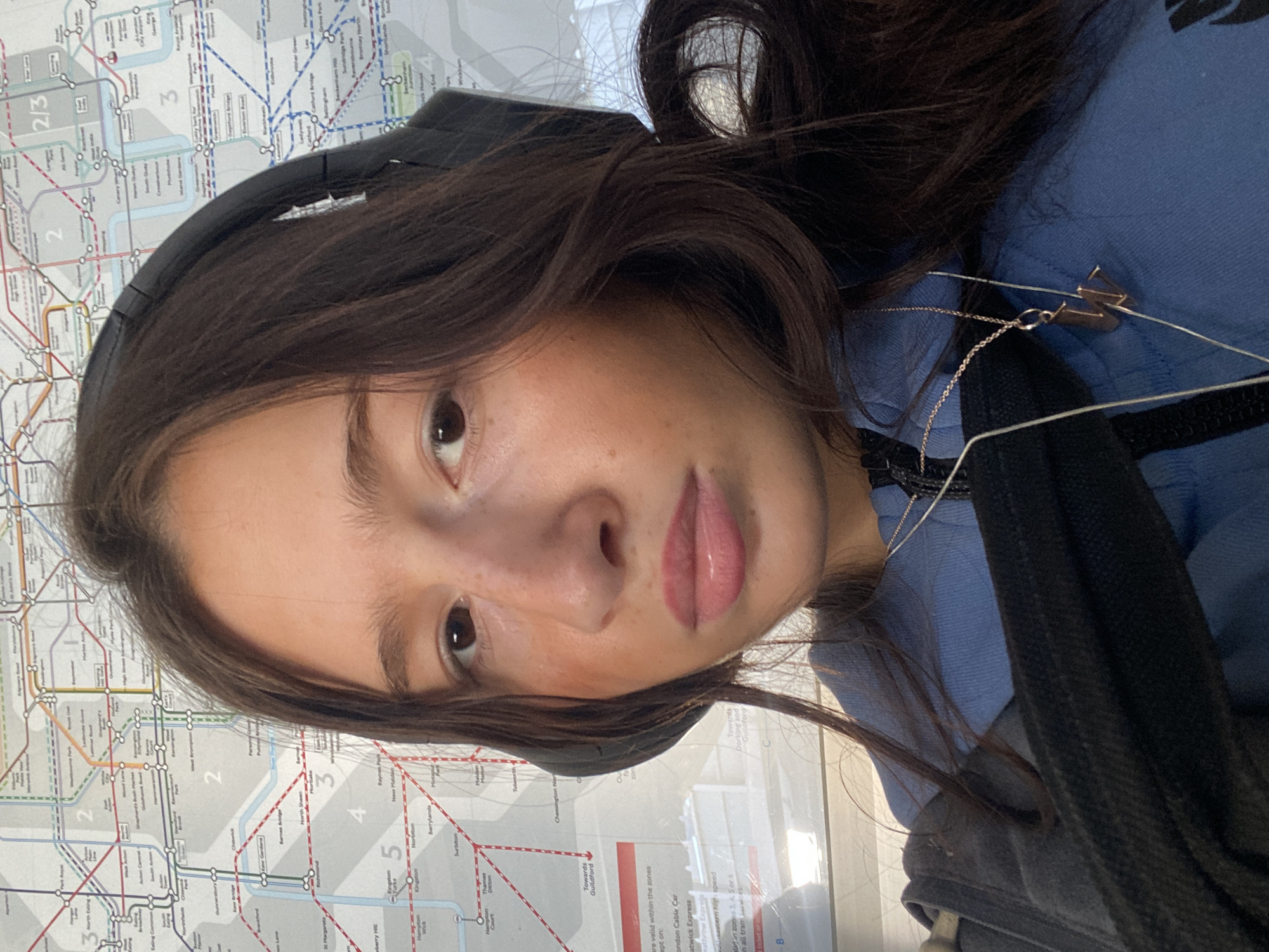 A young person, likely female, is featured in a close-up selfie, positioned vertically against a background of a public transport map. She has long, dark brown hair, which frames her face and partially obscures a black over-ear headphone on her left side. Her expression is neutral as she looks directly at the camera. She is wearing a light blue hooded top or sweatshirt, and a delicate silver-colored chain necklace with a small, gold-toned "M" pendant is visible on her right shoulder. White earphone cables are also draped around her neck and shoulder. A black backpack strap is visible on her right.
The background is a detailed map, which is clearly identifiable as a London transport map due to visible text such as "London Cable Car," "Gatwick Express," "Heathrow Express," "Towards Guildford," and station names including "Kingston," "Richmond," "Canary Wharf," and "Sutton." Fare zones "2/3," "2," "3," "4," "5," and "6" are also visible. The setting appears to be indoors, likely on public transport or in a station environment, with bright, diffused lighting suggesting daytime. The activity is the person taking a selfie, possibly while traveling or waiting.
