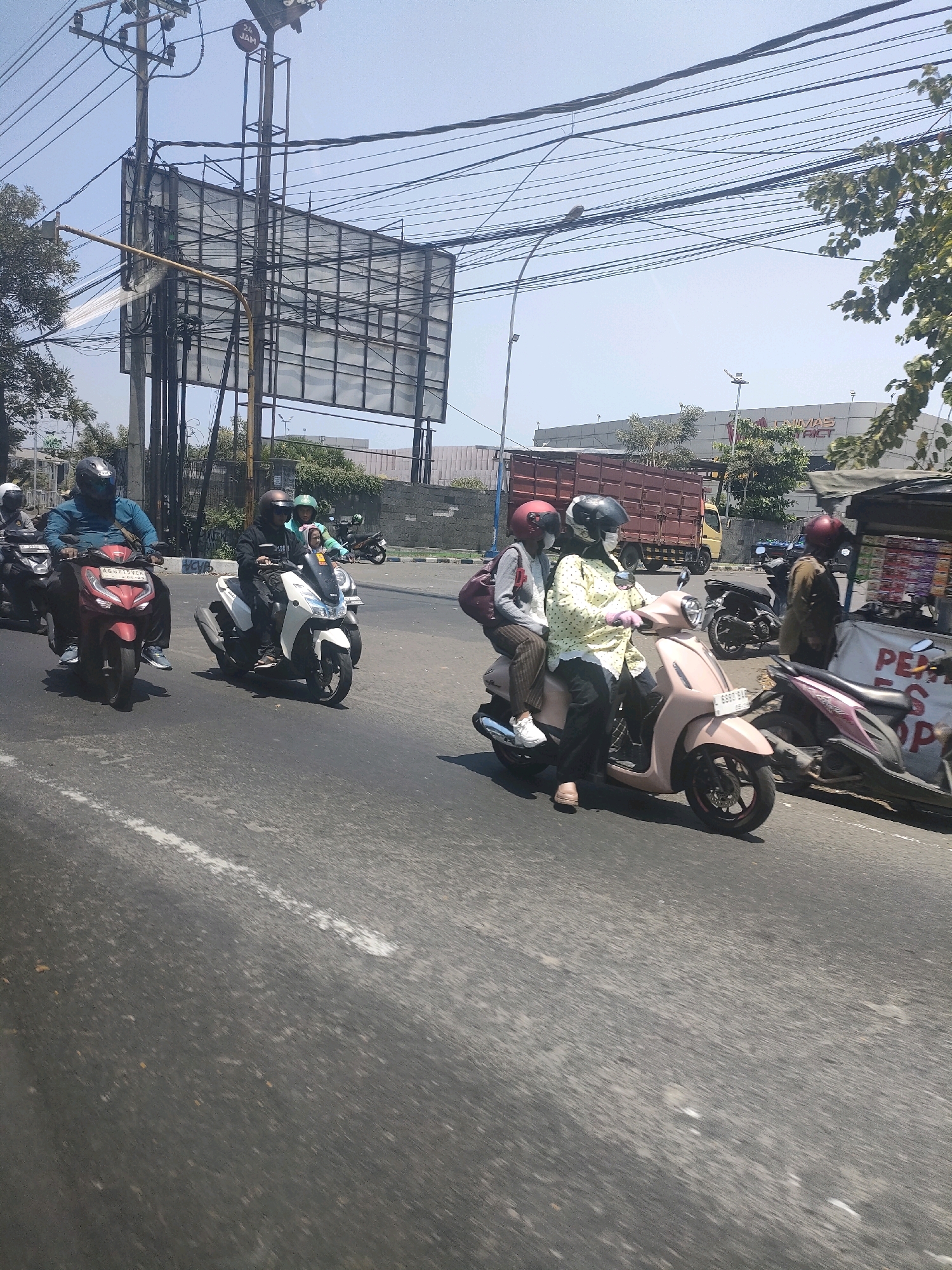 A group of motorcyclists are traveling on a street in Waru, Indonesia. The road appears to be asphalt with a white lane marker. The sky is clear and blue, indicating a sunny day.
On the left side of the road, a man wearing a blue shirt and a black helmet rides a red motorcycle. Behind him, another motorcyclist is partially visible.
In the center of the image, a white motorcycle carries two people. The rider wears a black jacket and a helmet, while the passenger behind them has a green helmet and appears to be a child. Further back, a person in a red helmet rides a dark motorcycle.
On the right side of the road, a pink scooter is ridden by a person in a yellow polka-dot shirt and a white face mask. They are wearing pink gloves and black pants. A passenger sits behind them, wearing a grey sweater and a red helmet. Adjacent to them, a stall is set up, possibly selling goods, with items stacked on shelves. A pink and black motorcycle is parked behind the stall. In the background, a large truck with a brown cargo container is visible, along with buildings and overhead power lines. A billboard structure stands prominently in the mid-ground on the left.