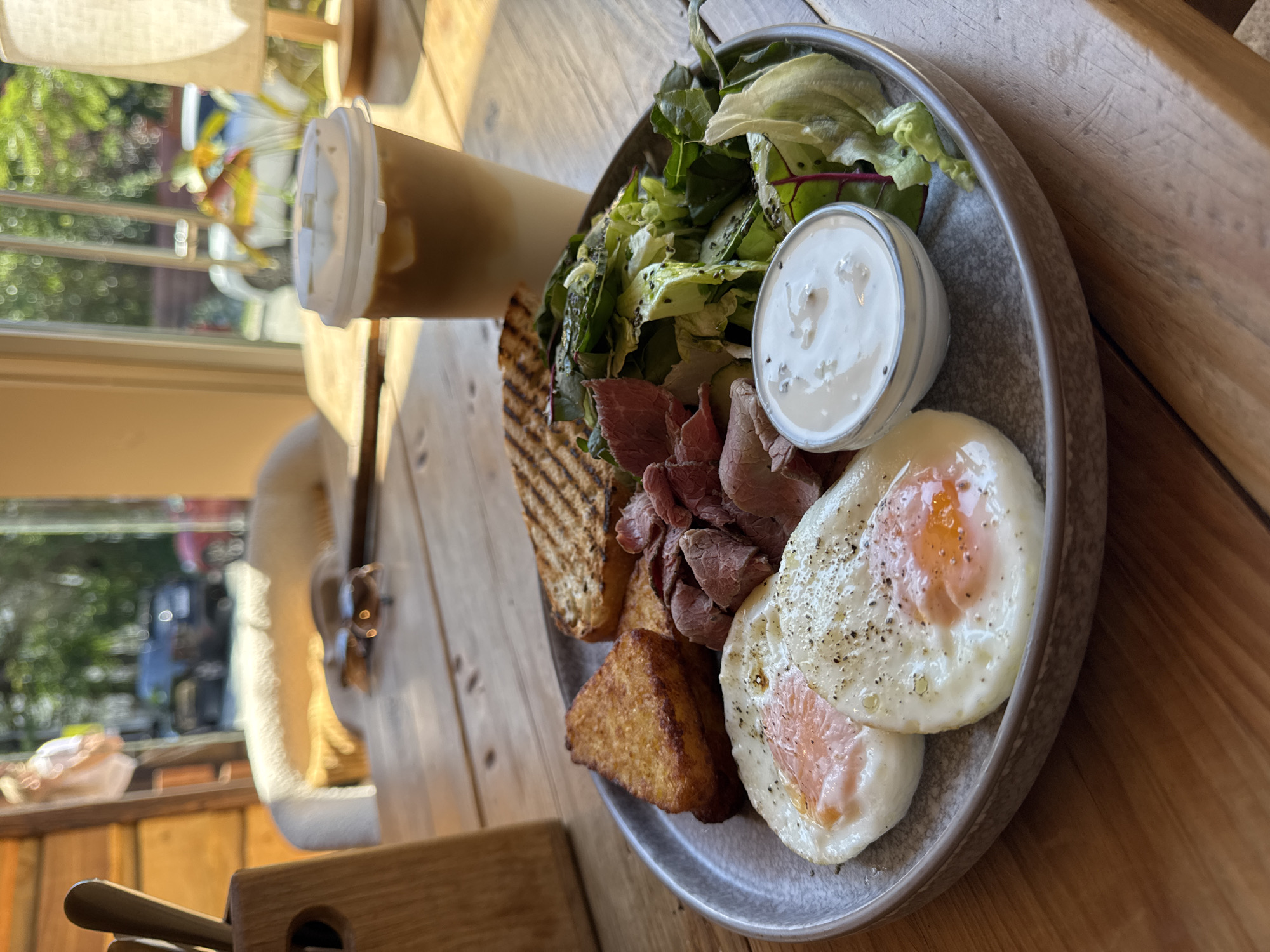 A close-up, eye-level, vertical shot showcases a breakfast plate on a rustic wooden table. The plate holds two fried eggs with visible yolks, seasoned with black pepper, a triangular hash brown, thin slices of roast beef, grilled toast, a small salad with leafy greens and sliced cucumber, and a ramekin of white sauce. To the right of the plate is a tall to-go cup with a white lid, containing a light brown beverage with ice. In the blurred background, sunlight streams through large windows, illuminating a chair with a fluffy white seat, a wooden utensil holder with cutlery, and a lamp with a beige shade. Outside the window, foliage and vehicles are visible, suggesting an outdoor or semi-outdoor dining setting. The overall scene depicts a breakfast meal in a cafe or restaurant environment, possibly in Krasnaya Polyana, Russia, given the ambient light and natural elements.