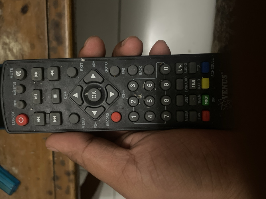A person's right hand holds a black remote control. The remote is a Venus brand and has buttons for various functions, including standby, info, subtitle, mute, record, menu, exit, volume, channel, goto, epg, back, media, usb, tv/radio, l/r audio, fav, timer, 16:9 ratio, ttx, dpi, 1080p, and schedule. The hand is holding the remote in front of a wooden surface. The background of the photo is a white wall. The scene appears to have been captured indoors. The coordinates provided correspond to a location in Cimenyan, Indonesia. It is unknown why the person is holding the remote, but there is no evidence of any other actions or interactions in the image.