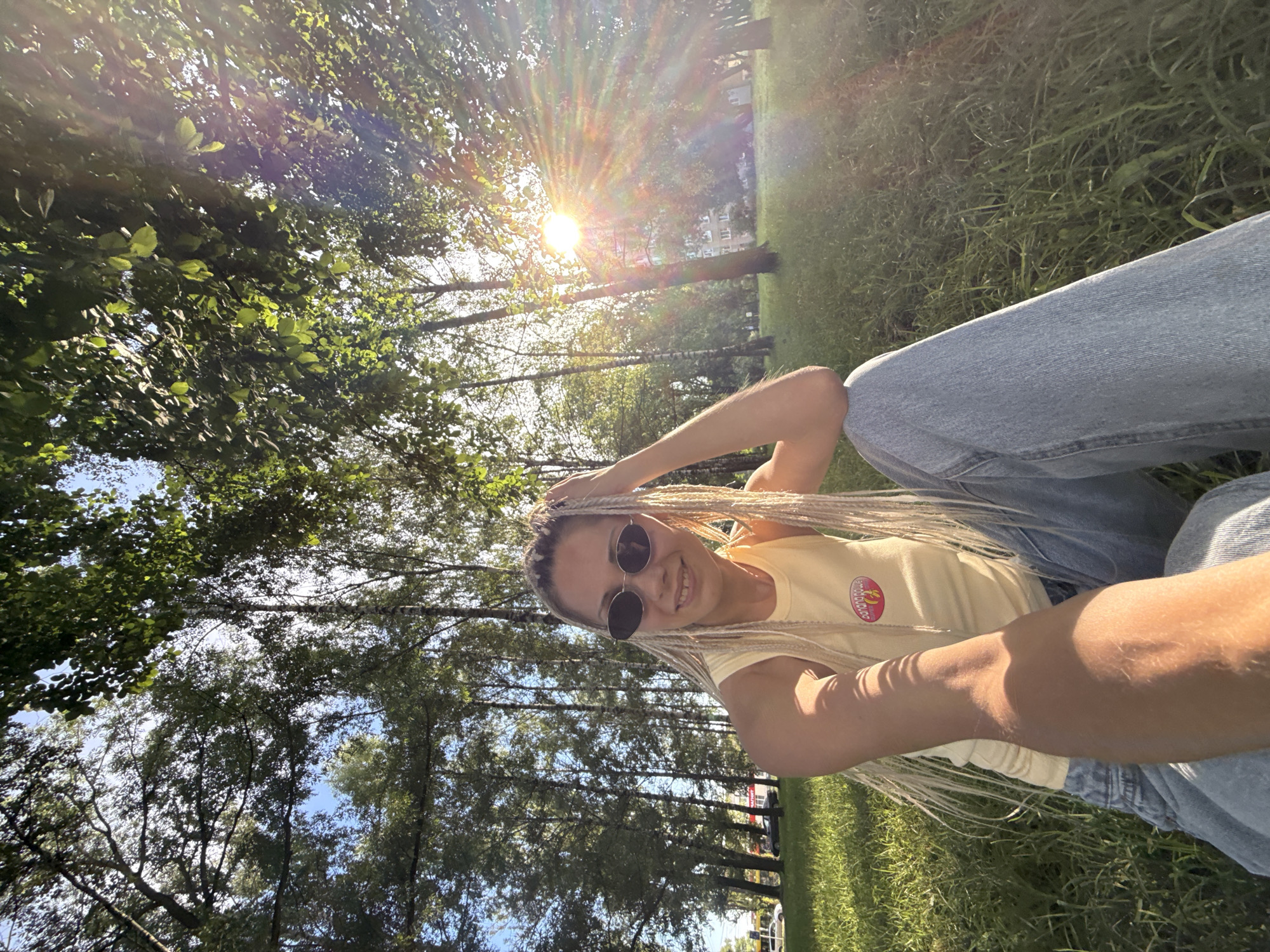 A young woman with blonde braids and sunglasses sits on a grassy lawn in a park, looking at the camera and smiling. The sun is shining brightly, creating lens flare. Trees with green leaves are visible in the background, along with a building and cars in the distance. The woman is wearing a yellow sleeveless shirt and blue jeans. The image appears to have been taken in the city of Zhukovo, Russia.