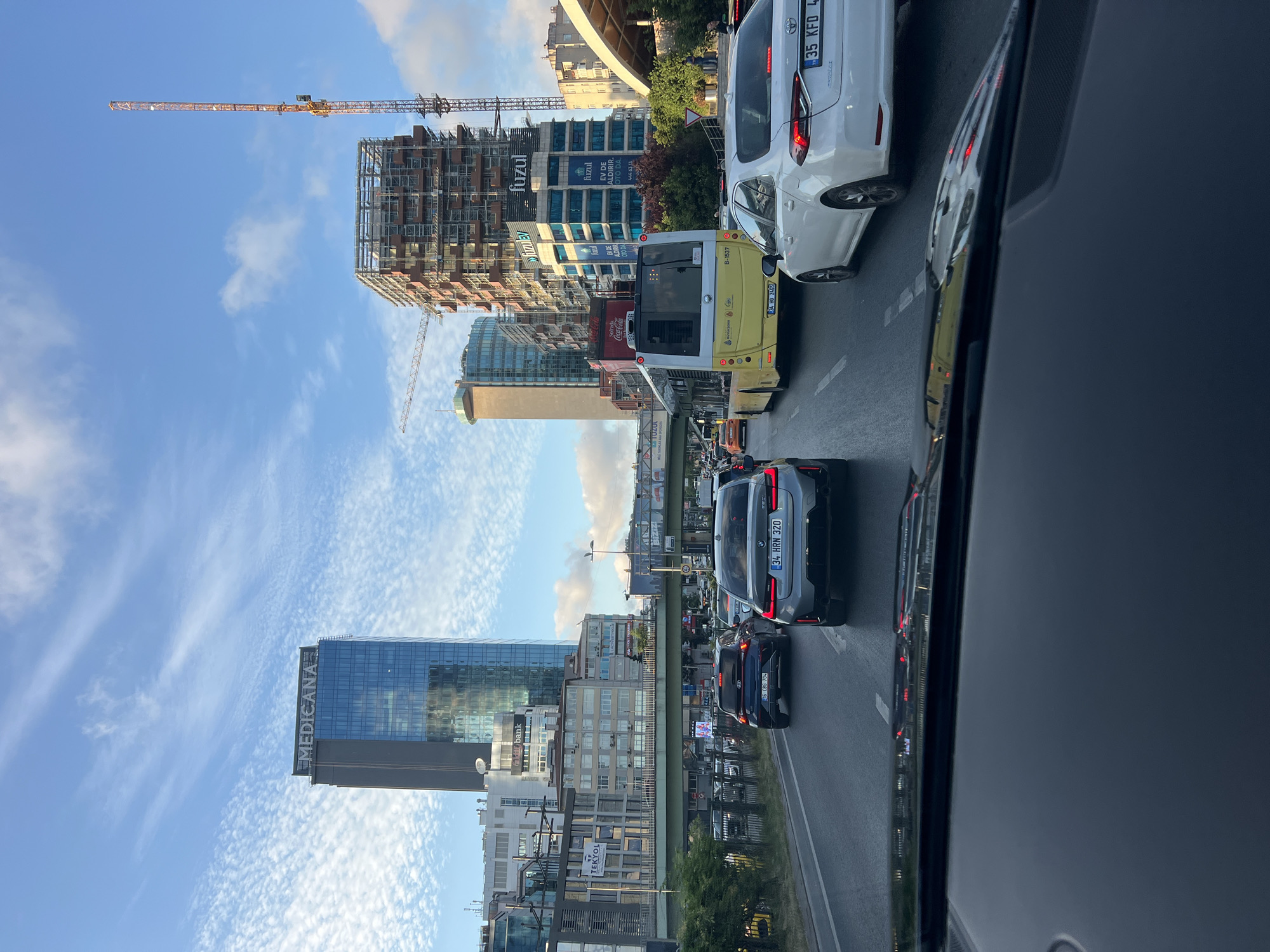This is a vertical, slightly tilted, eye-level shot of a busy street scene in Beşiktaş, Turkey. The image captures a traffic jam with several cars and a yellow bus stopped on a multi-lane road. The architecture is modern, with several tall buildings visible, including one with "MEDICANA" prominently displayed and another under construction with a large crane. Other visible text includes "TEKYOL," "fuzul," "Sofrada," and "Coca-Cola," suggesting various businesses and advertisements. The sky is a bright blue with wispy white clouds, indicating it's daytime, likely late afternoon or early evening given the lighting. The overall atmosphere appears to be one of urban hustle and bustle, characteristic of a major city.