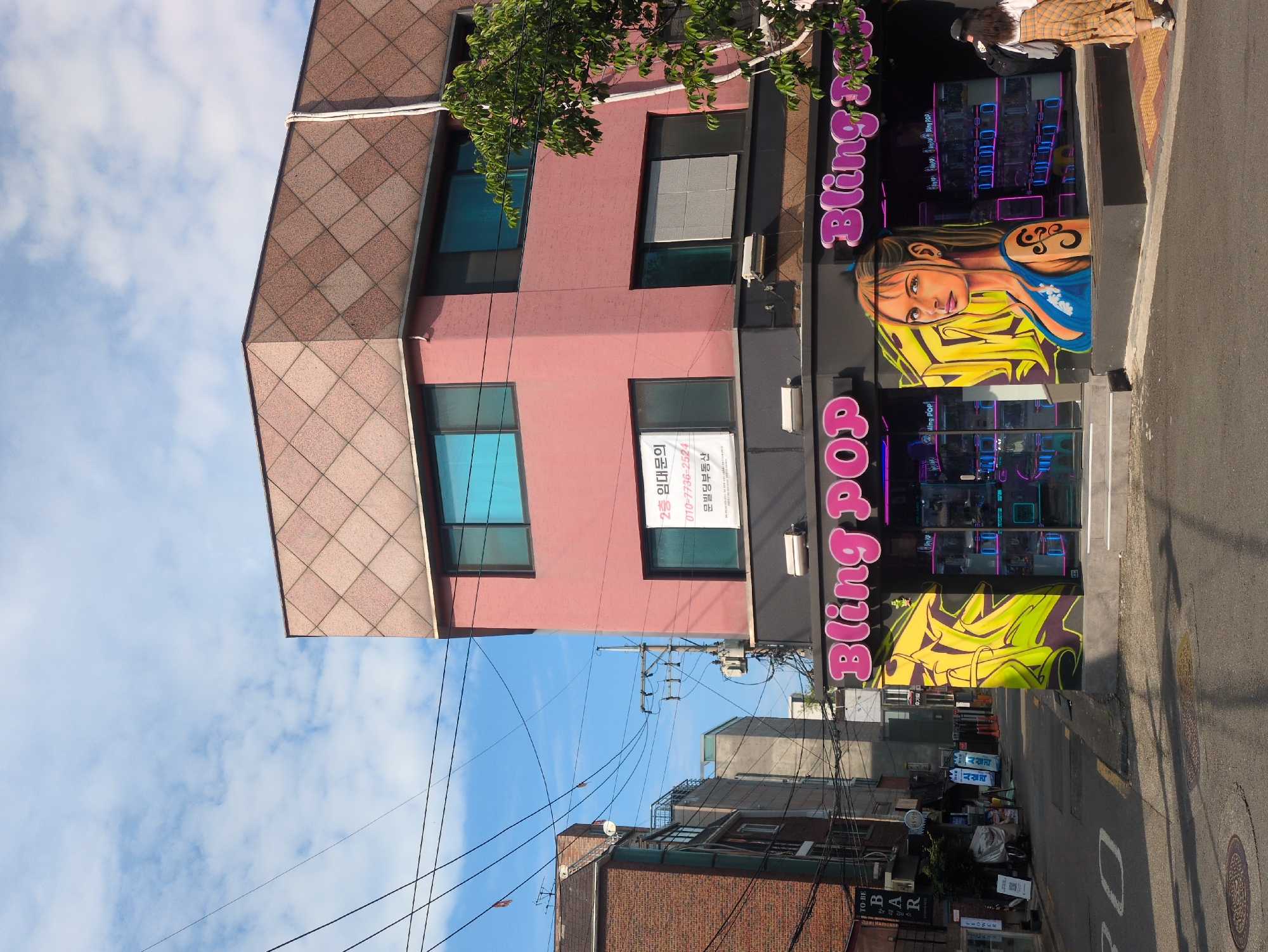 Here is a description of the image:
The image captures a street scene in Seoul, South Korea, featuring a commercial building. The building has a distinctive architectural design, with a portion of its facade covered in brown-tiled square patterns and another part painted in a muted pink. The building's windows are rectangular, framed in black, and reflect a blue sky. A sign in Korean is visible on the building's pink section.
The storefront of the building displays a vibrant and eye-catching mural and signage for "Bling Pop," with a stylized image of a girl with blonde hair, a yellow-green abstract graphic, and a large neon-style "Bling Pop" inscription.
The street is relatively narrow, with other buildings visible in the background. Power lines crisscross the sky above the buildings, adding to the urban environment. Street signs, trash bags, and discarded items dot the sidewalk, suggesting a busy urban environment. A woman is present in the image standing in front of the storefront.