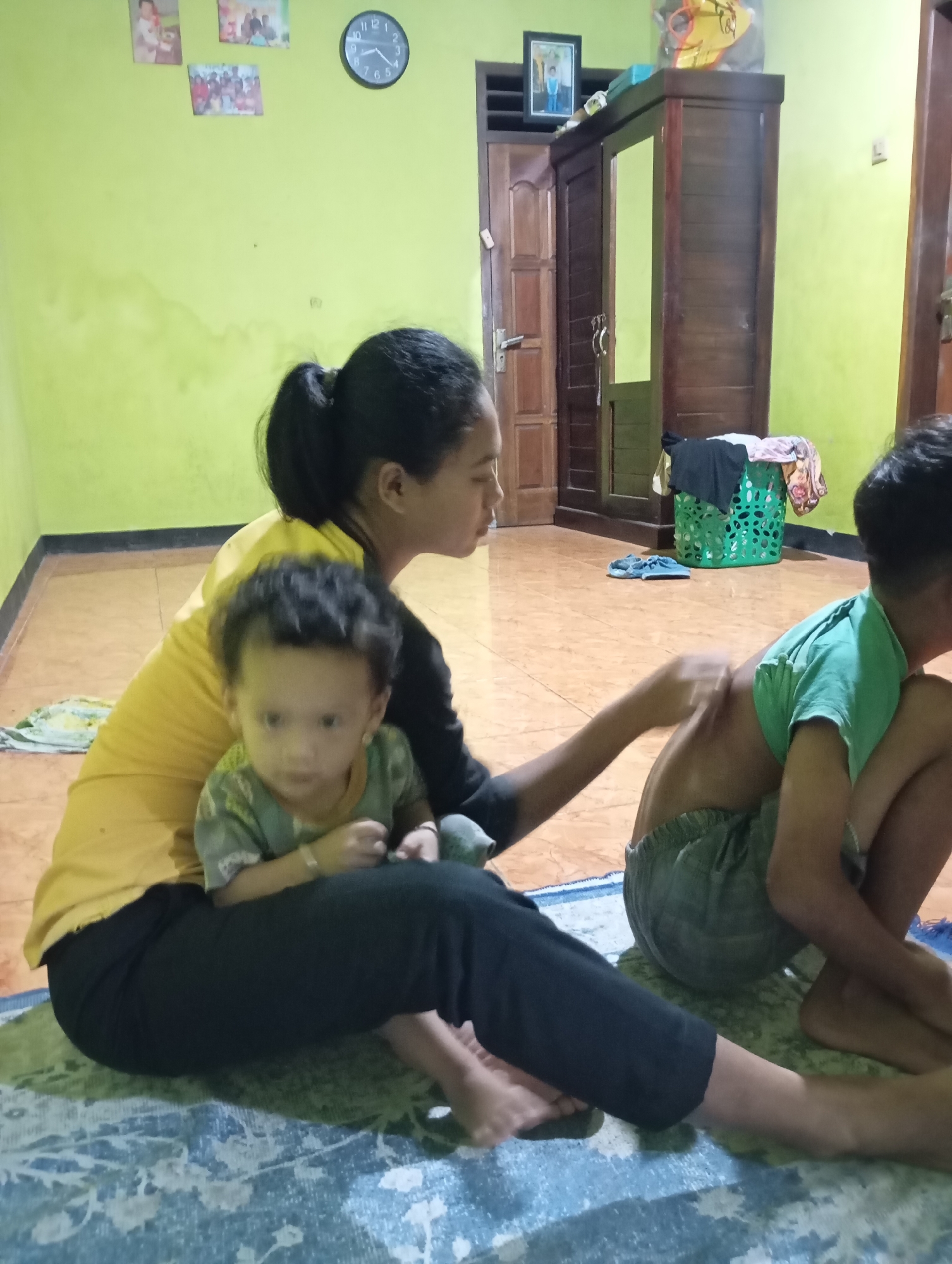 The image depicts an indoor scene, likely a home in Ketepung, Indonesia, based on the provided context. In the foreground, a woman is sitting on a blue and green patterned rug, with a young child in her lap. The woman wears a yellow shirt and black pants, and her hair is pulled back in a ponytail. The child, facing the viewer, is wearing a green and grey patterned shirt and appears to be looking towards the camera. To the right of the woman and child, a second child, a boy wearing a green t-shirt and grey shorts, is seated, and the woman's hand is touching his back, suggesting an interaction such as a massage or gentle touch.
The background reveals a room with lime green walls. On the wall to the left, there are three framed pictures and a round clock with Roman numerals. In the background to the right, a large, dark wooden wardrobe with double doors and a mirror is visible. A green plastic laundry basket filled with clothes is placed near the wardrobe. The floor is tiled with a warm, orange-brown tone. The overall impression is of a domestic environment.