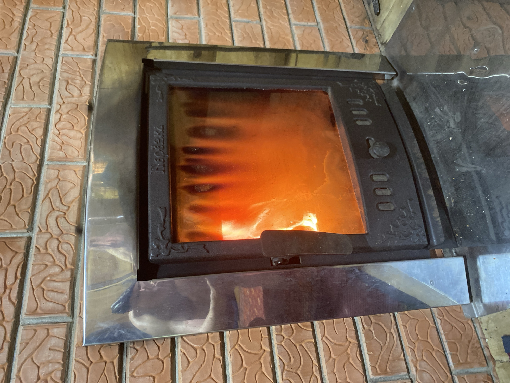 The media file displays a close-up of a wood-burning fireplace insert with an active fire, set into a tiled wall.
The main subject is a modern fireplace insert with a large, rectangular glass door, behind which bright orange and yellow flames are visible. The fire appears vigorous, casting a warm glow. Dark, vertical streaks, likely soot or residue, are present on the inside surface of the glass door. The fireplace door has a dark-colored, possibly cast iron, frame with some decorative patterns on the right side and a handle at the bottom. Along the left vertical edge of the door, the Cyrillic text "Бердезъ" (Berdez') is inscribed.
The fireplace insert is surrounded by a polished, silver-toned metallic trim that reflects parts of the room. This trim transitions smoothly into the surrounding wall.
The wall itself is covered with rectangular, light brown or terracotta-colored tiles, arranged in a staggered, brick-like pattern. Each tile has a distinctive, raised, wavy, or organic relief texture across its surface, adding a decorative element to the setting.
The scene depicts an indoor environment, with the fireplace being the focal point, suggesting warmth and comfort. The location context provided is Гаревая, Russia. There are no people visible in the image, and the time of day is not discernible.
