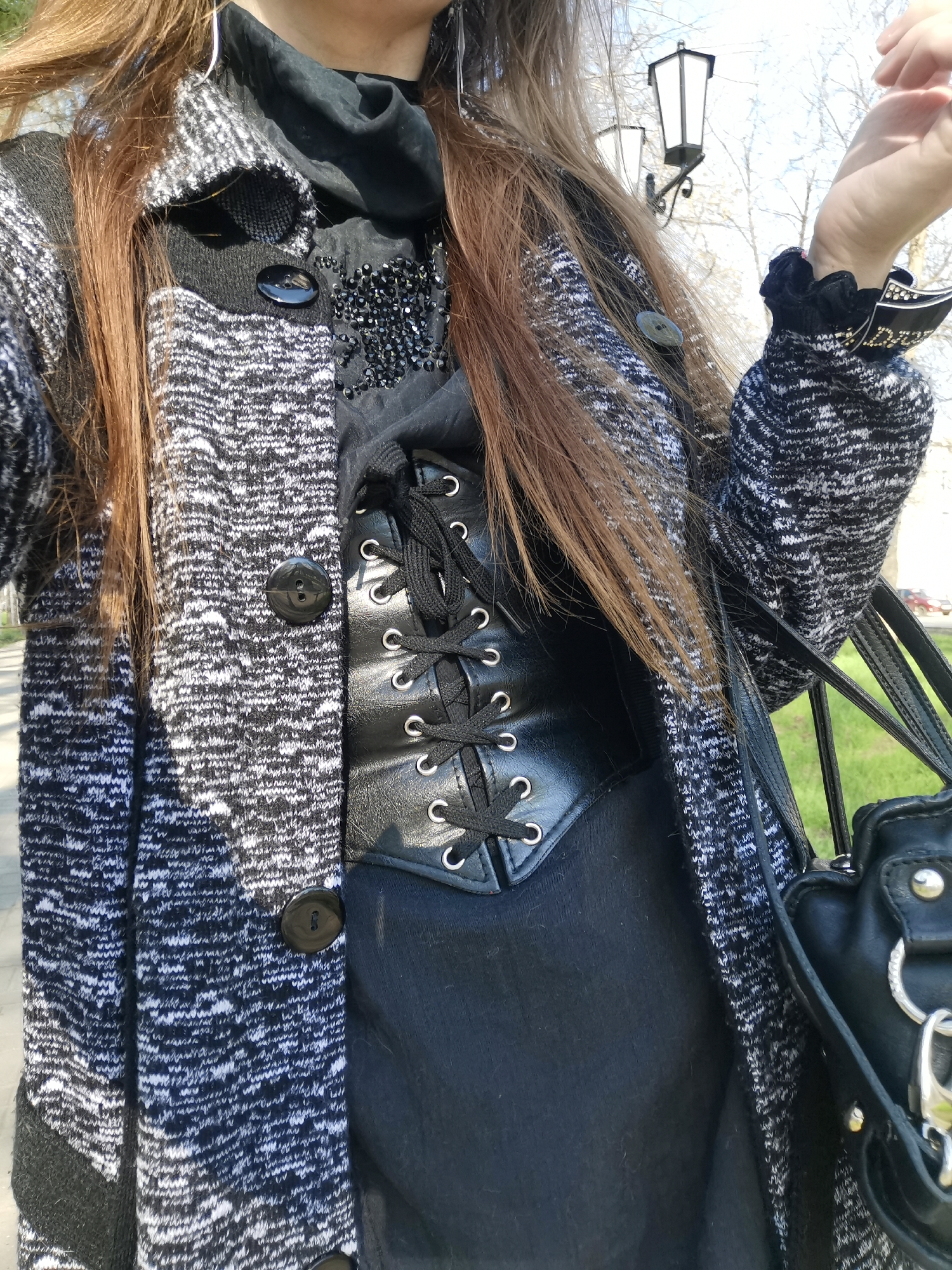 A full shot captures an individual on a cobblestone sidewalk in an urban area of Unknown, Unknown. The individual dons a black and white patterned cardigan, secured with round, black buttons. Underneath, a black, lace-up corset is layered over a dark, beaded top with a sheer collar. The person also wears a silver, drop-style earring. The person also carries a black leather handbag, dangling from their wrist. A black vintage-style lamp post and bare-branched trees form the backdrop.
