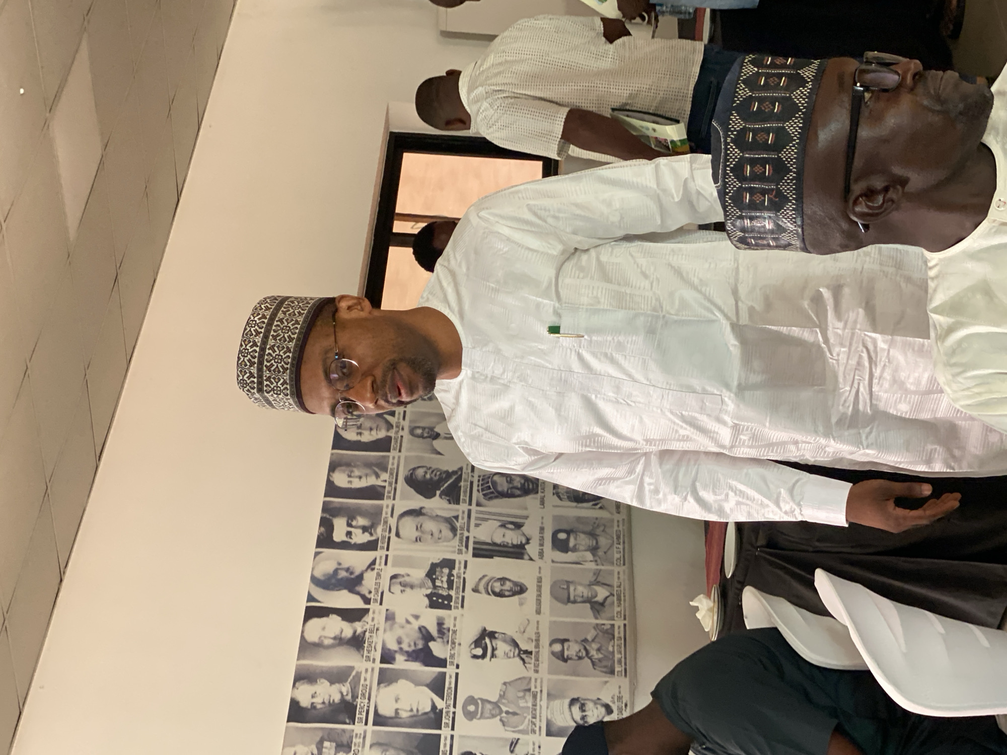 Here's a description of the image:
**Location:** Kwarau, Nigeria (assumed, based on context)
**People:** Several men are visible. Two are in the foreground. One wears a white garment and a patterned cap, and glasses. Another has a similarly patterned cap, and a white garment, but is seen from the back. Other individuals are partially visible in the background, at least one wearing a white shirt.
**Objects:** A wall has a collage of monochrome portrait photos of men, each labeled with text, including the names "SIR PERCY GIROUD," "SIR HESKETH BELL," "SIR CHARLES TEMPLE," "SIR JOHN PATTERSON," "ERIC THOMPSTONE," "SIR GAWAIN BELL," "ABBA LABAL KAITA," "CDABOERE," "COLLABIAL JAFARU SA," "COL HAMMED ALI," "COLUF AHMED," a pen is attached to the pocket of one of the individual's white garment, white plastic chairs, plates with napkins are visible at the table.
**Environment:** An indoor setting, possibly an office or meeting room. The lighting appears to be artificial.
**Actions/Interactions:** The people seem to be at a meeting or gathering. No specific actions are clearly visible.
