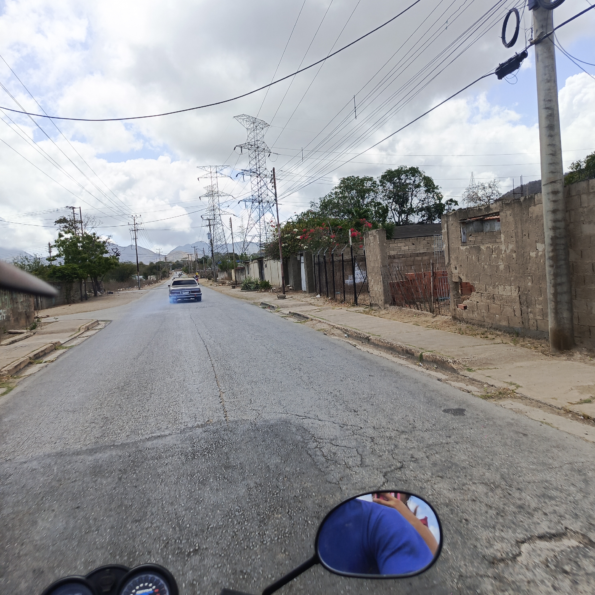 The image shows a street scene in Juan Griego, Venezuela. The viewpoint is from a moving motorcycle, as evidenced by the speedometer and rearview mirror in the foreground. A blue car is driving ahead on the road, emitting blue smoke from its exhaust. The road is paved but cracked. On the right side of the road is a sidewalk and a wall, partly made of cinder blocks, with sections of barbed wire. Utility poles and high-tension power lines are visible above. Mountains can be seen in the distance under a cloudy sky.