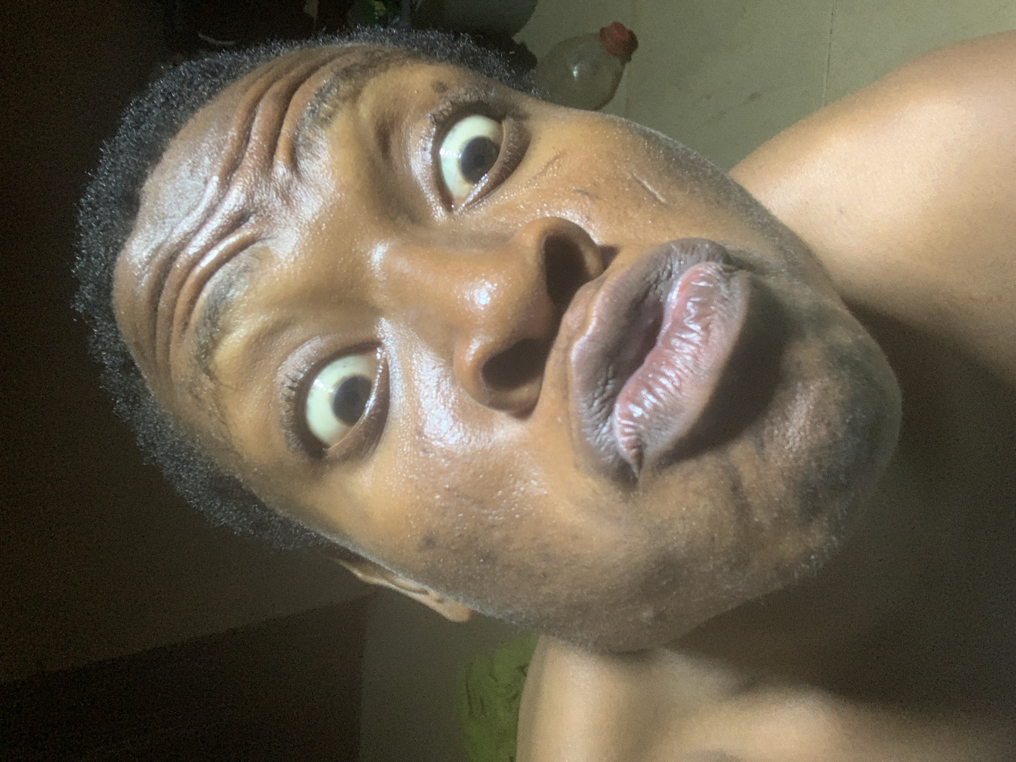 A close-up, angled shot of a dark-skinned individual with eyes wide open and lips pursed, positioned in an indoor environment. The location is Unknown, Unknown. The person's face is shiny, possibly due to perspiration or oil. Details include short, dark hair and visible wrinkles on the forehead. A plastic bottle is partially visible in the blurred background. The person's shoulder is also partly in the frame.