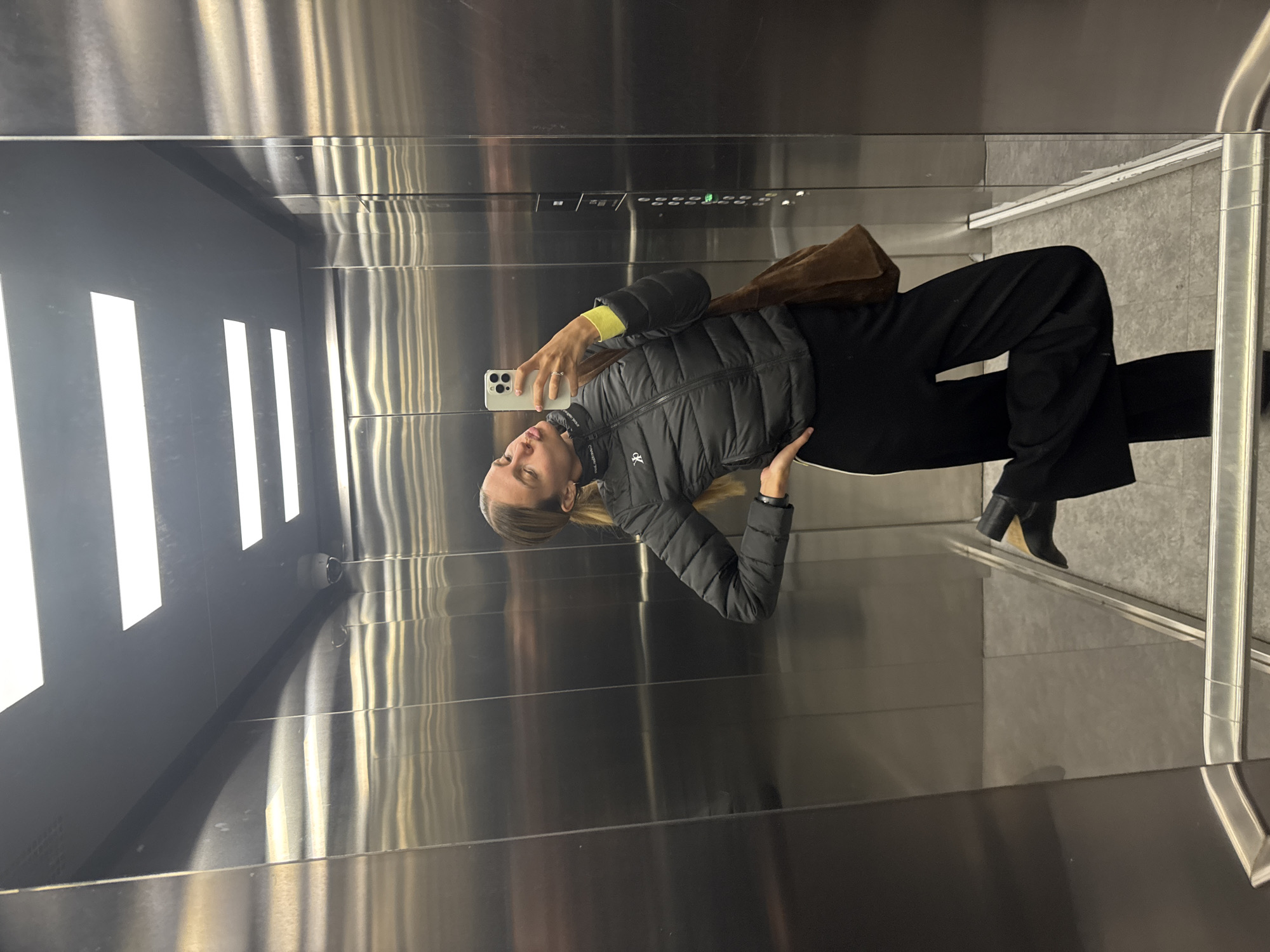 A woman is taking a selfie in an elevator. She is wearing a black puffer jacket, black pants, and black heeled boots. She is holding a smartphone in her left hand. A brown bag is hanging from her shoulder. The elevator has stainless steel walls and fluorescent lighting. The elevator control panel is visible. The location is unknown.