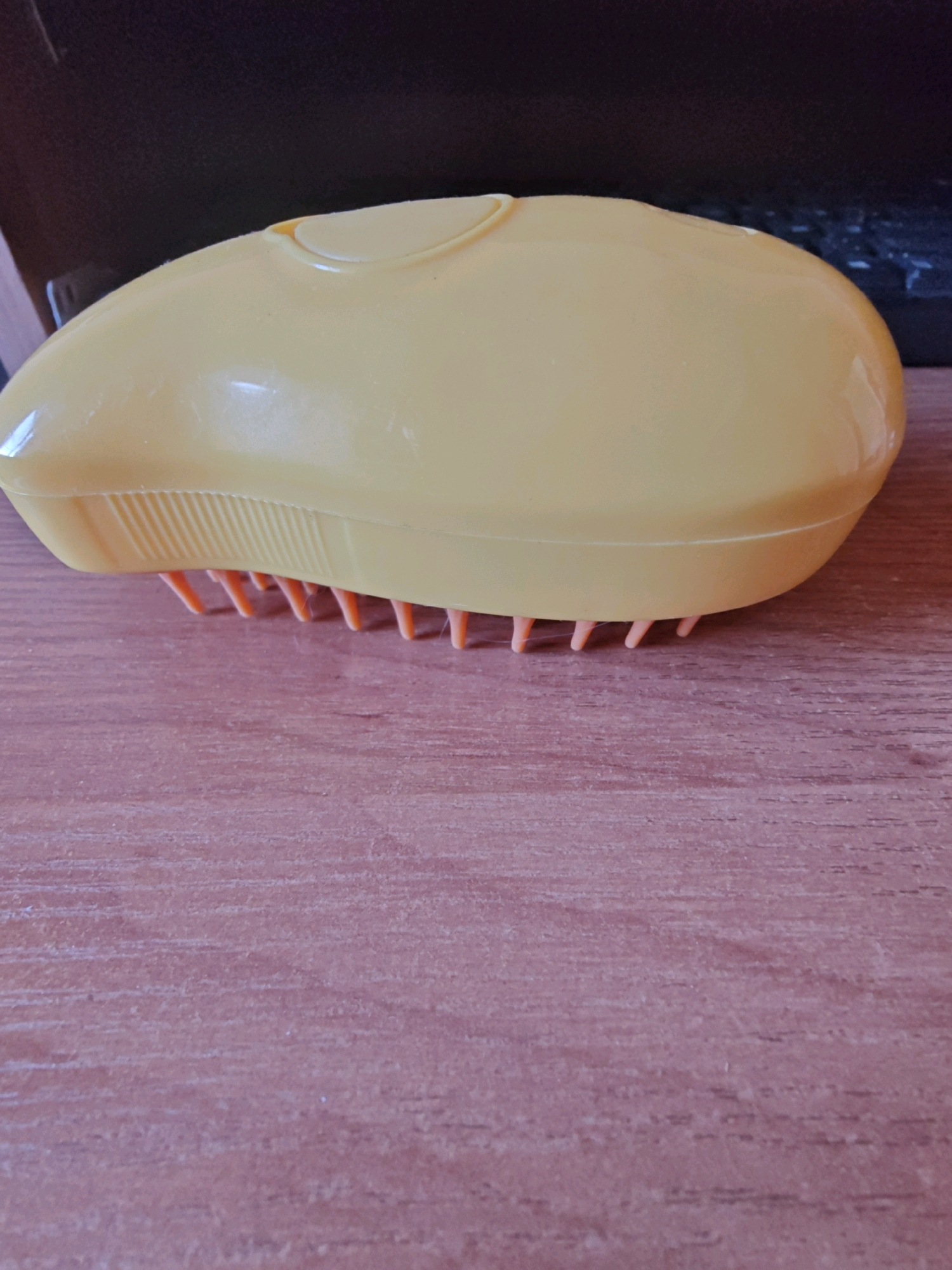 A yellow, oval-shaped brush with orange bristles rests on a wooden surface. The brush has a raised, curved section on its top. The surface it sits on appears to be a table or desk with a wood grain pattern. The background is dark and out of focus, suggesting an indoor setting. The image lacks any contextual clues to determine its location within Rubtsovsk, Russia.